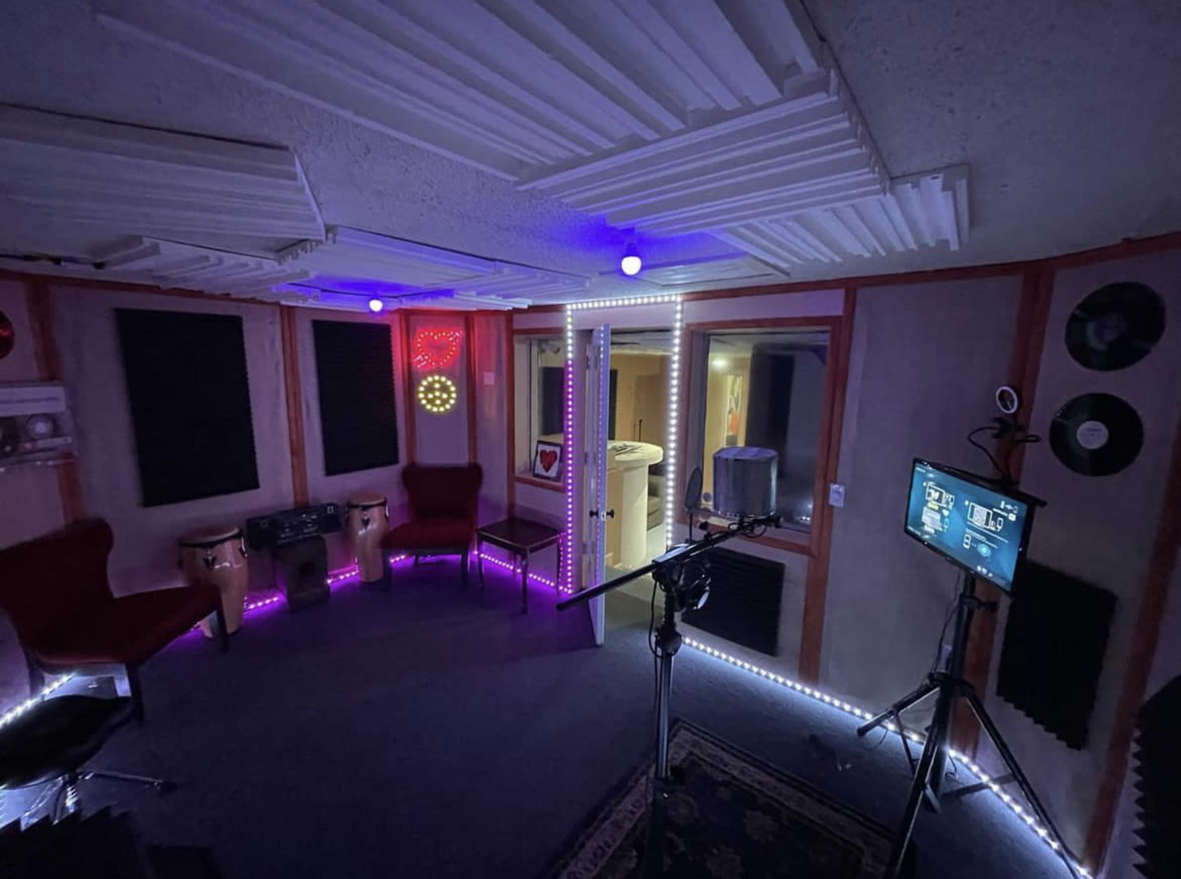 A recording studio featuring soundproof walls, two red chairs, a microphone on a stand, and colorful LED lighting.