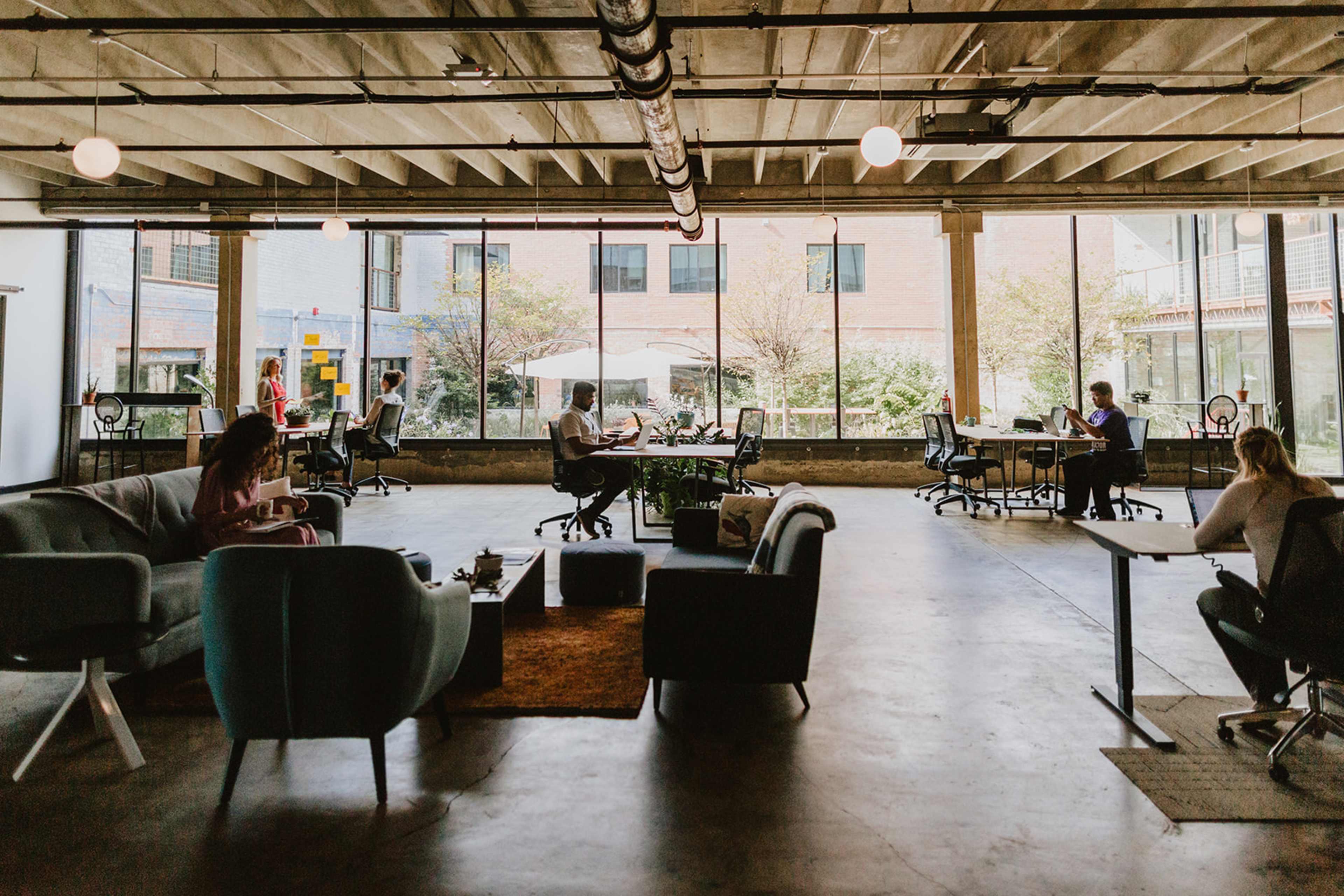 The image shows a modern co-working space with large windows, various seating areas, and individuals working at desks and sofas.