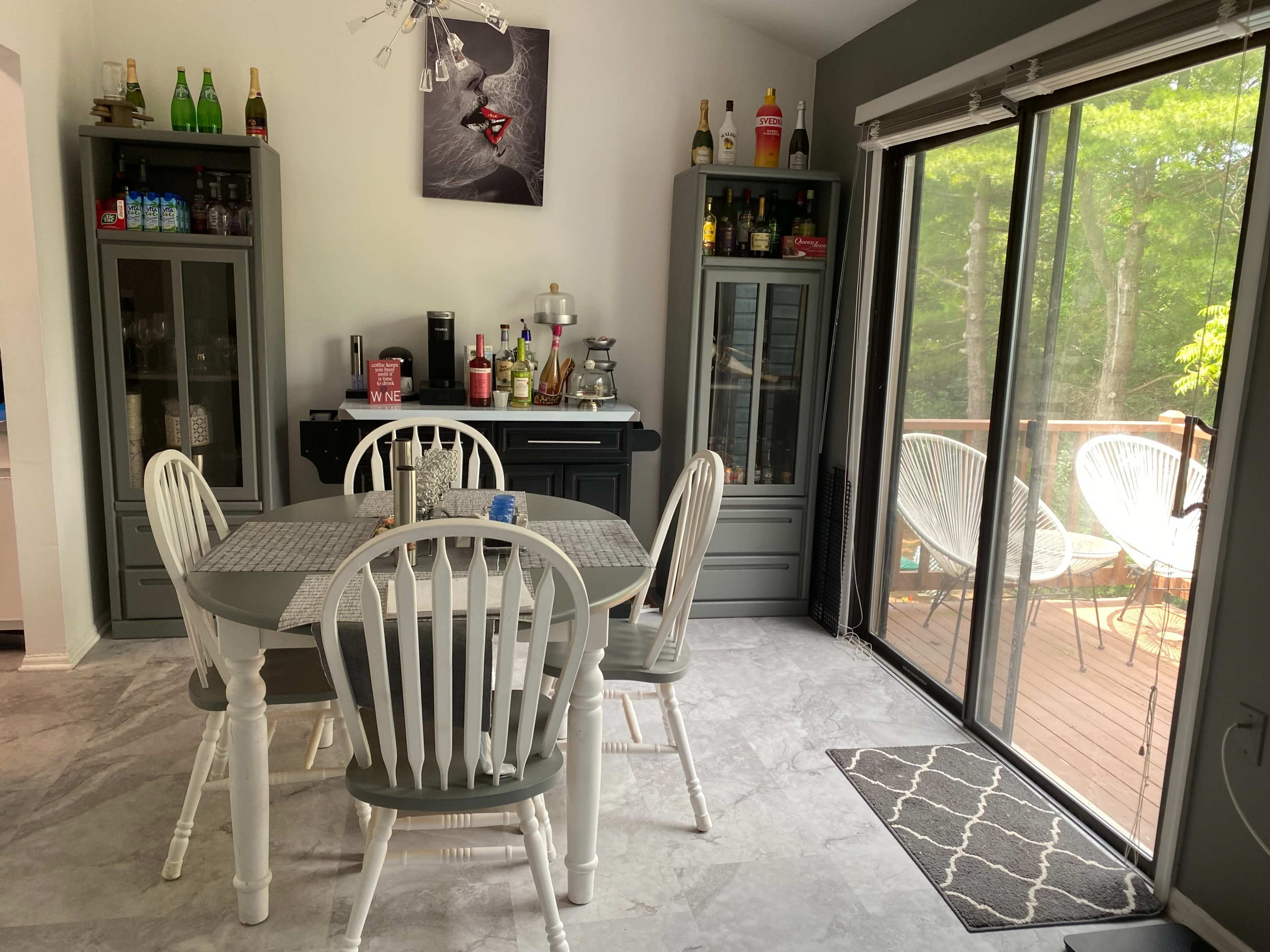 A dining area with a white table and four chairs, flanked by two cabinets filled with bottles, and a large sliding glass door leading to a deck outside.