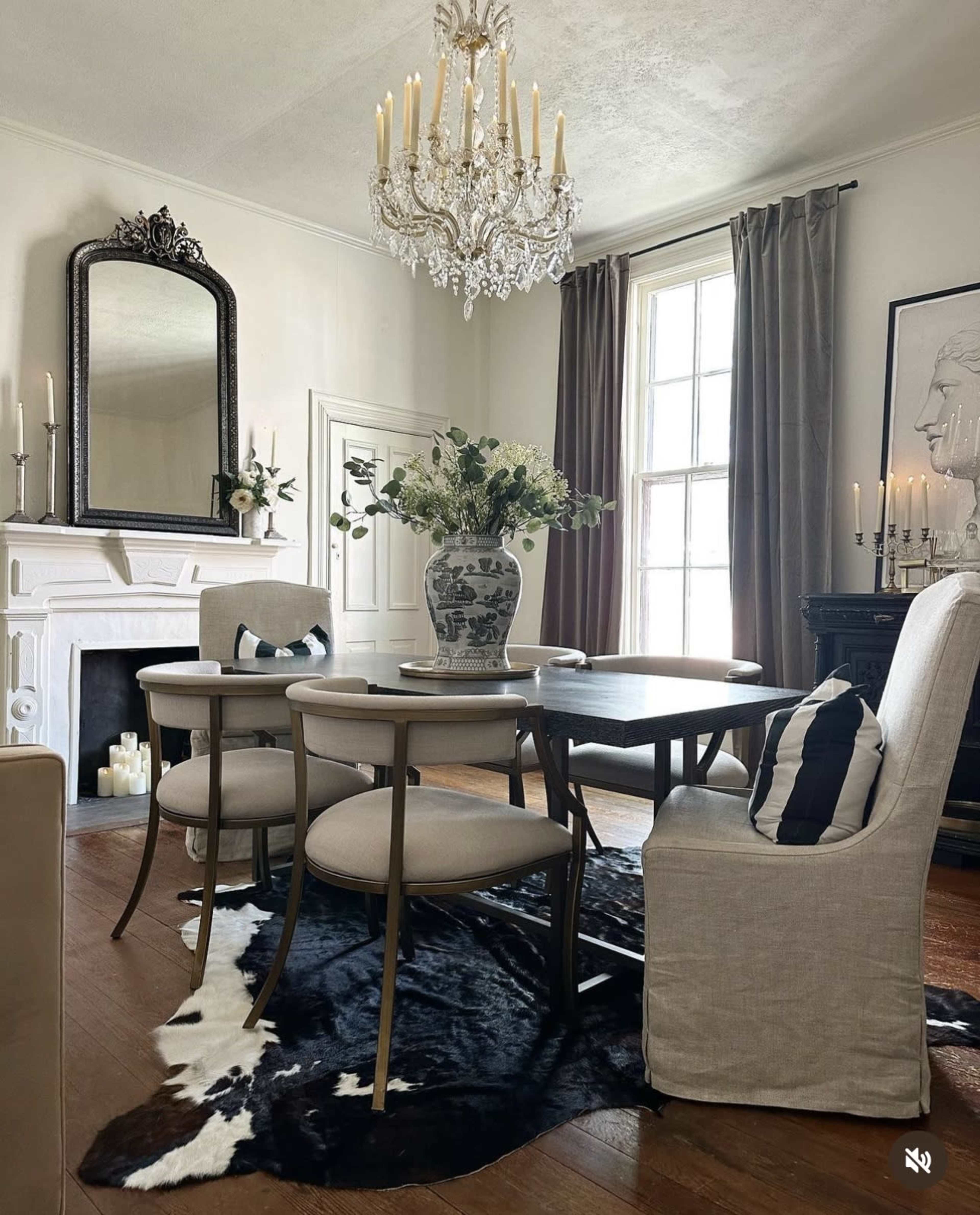 A dining room features a wooden table surrounded by upholstered chairs, a large vase with flowers on the table, and a chandelier above, with a decorative fireplace and art on the walls.