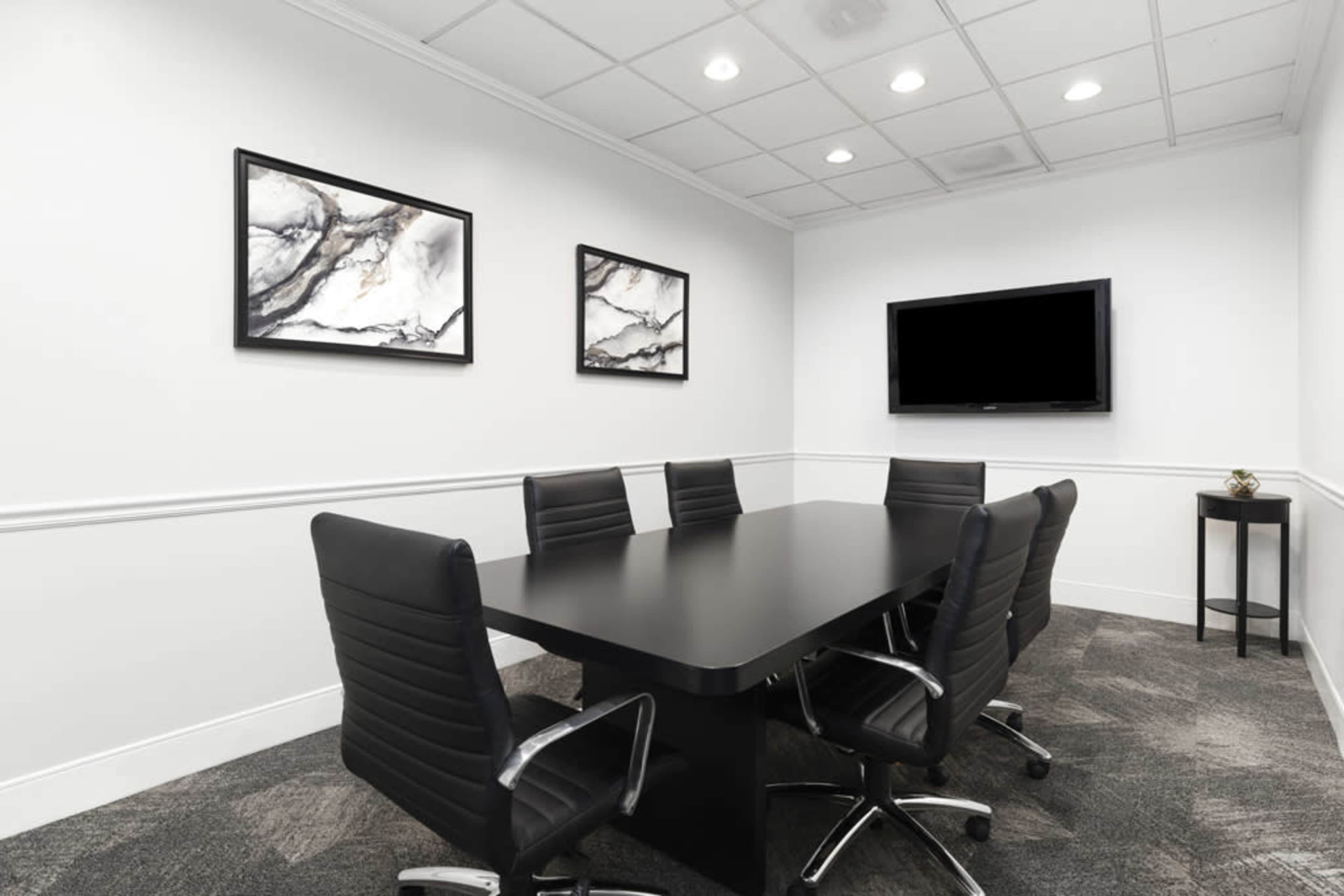 A modern conference room features a long black table surrounded by six leather chairs, with abstract art on the walls and a television mounted in front.