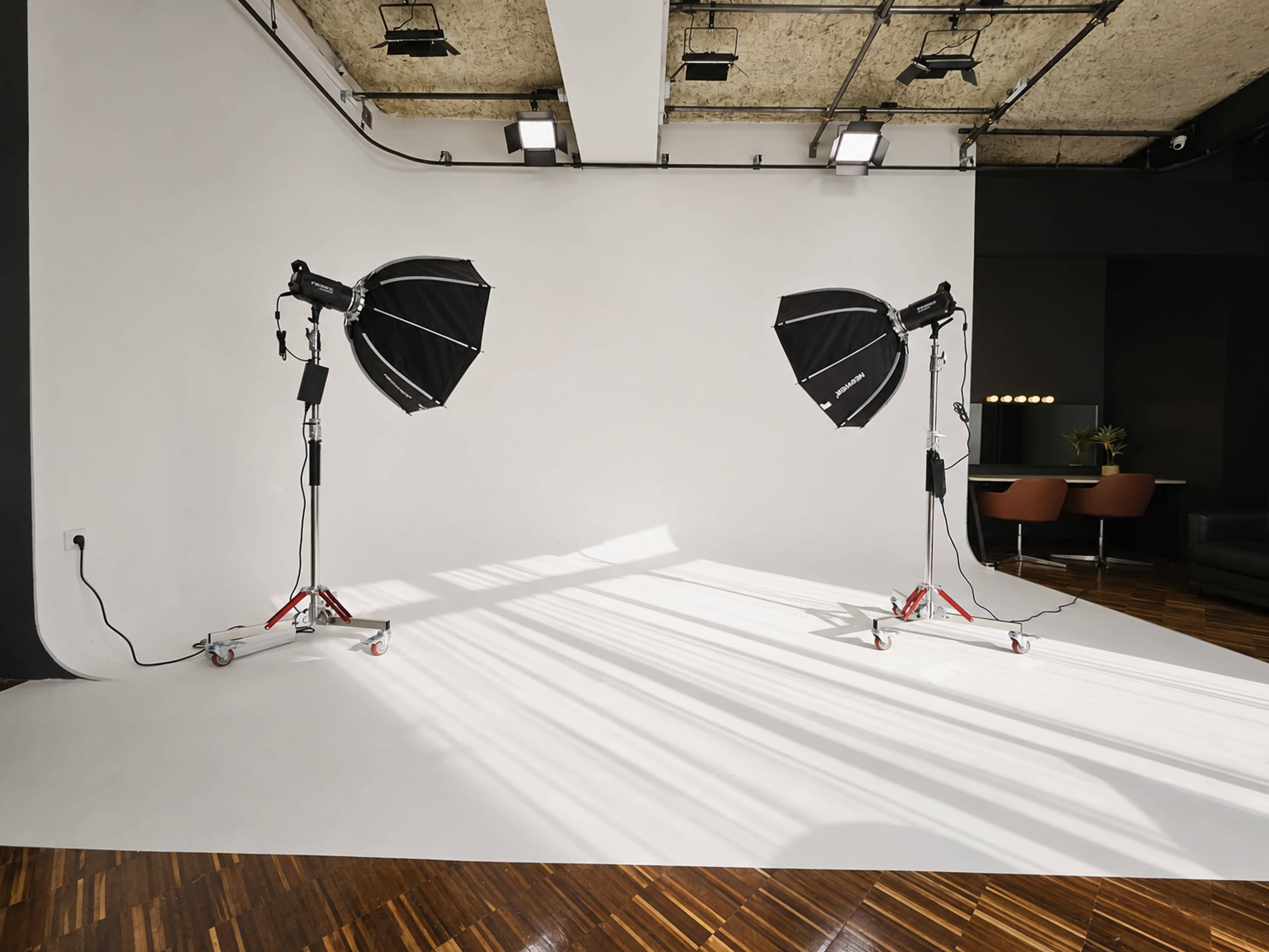 The image shows a photography studio with two large softbox lights positioned on either side of a blank white backdrop, casting shadows on the wooden floor.