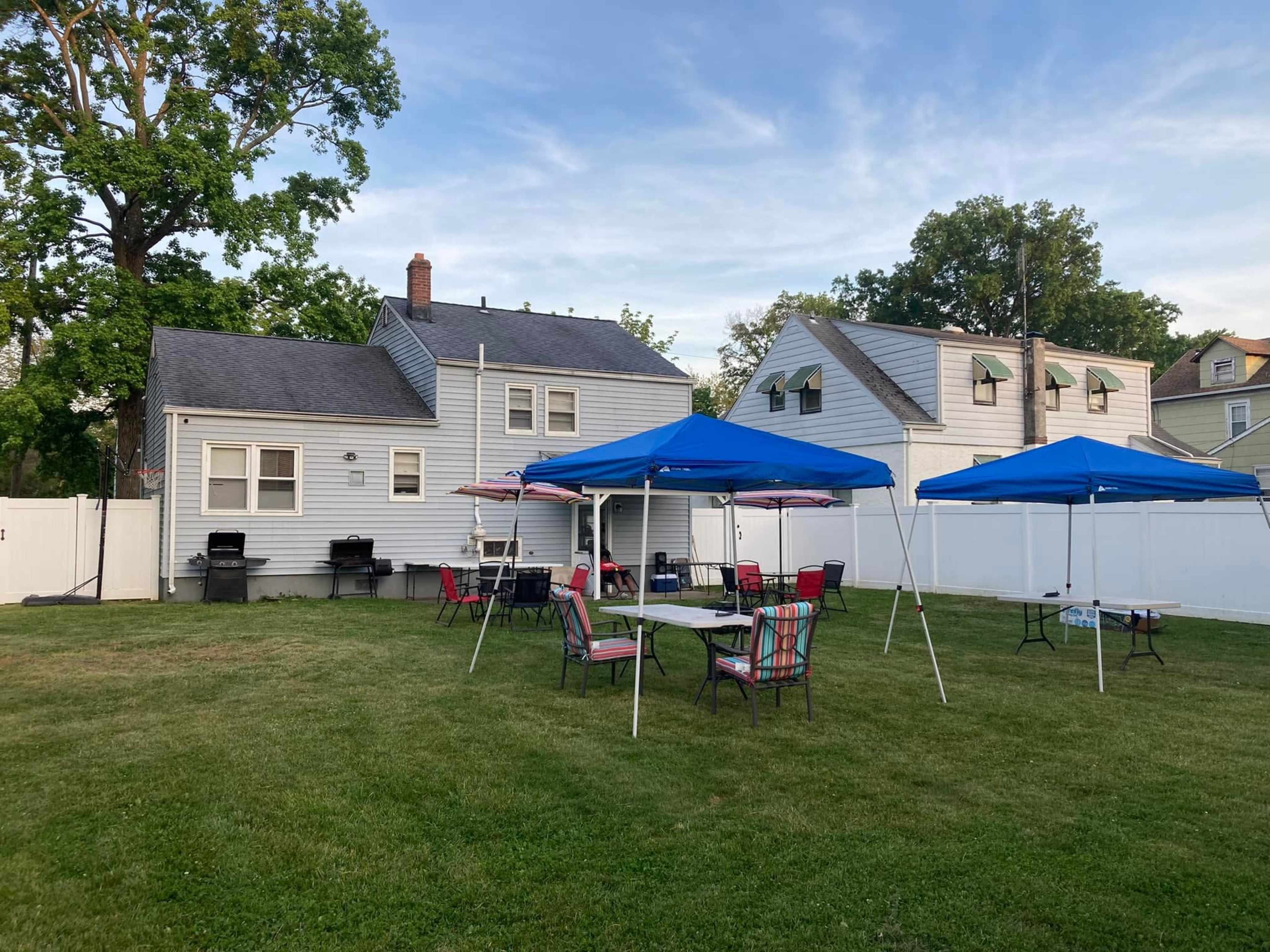 A grassy backyard features several blue tents, tables, and chairs set up near a white fence and two houses.