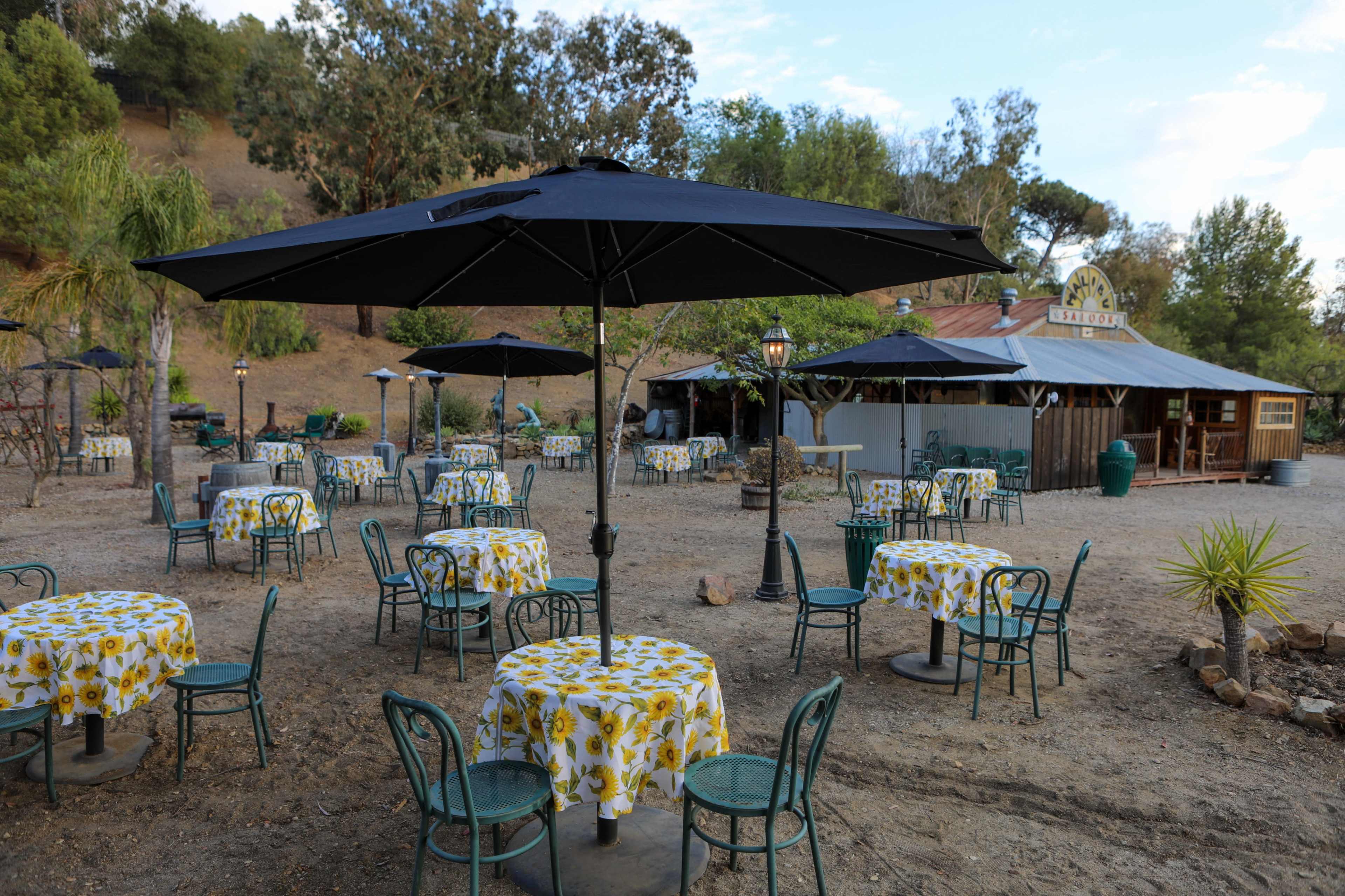 The scene shows an outdoor dining area with several tables covered in yellow floral tablecloths, black umbrellas, and a rustic building in the background surrounded by trees.
