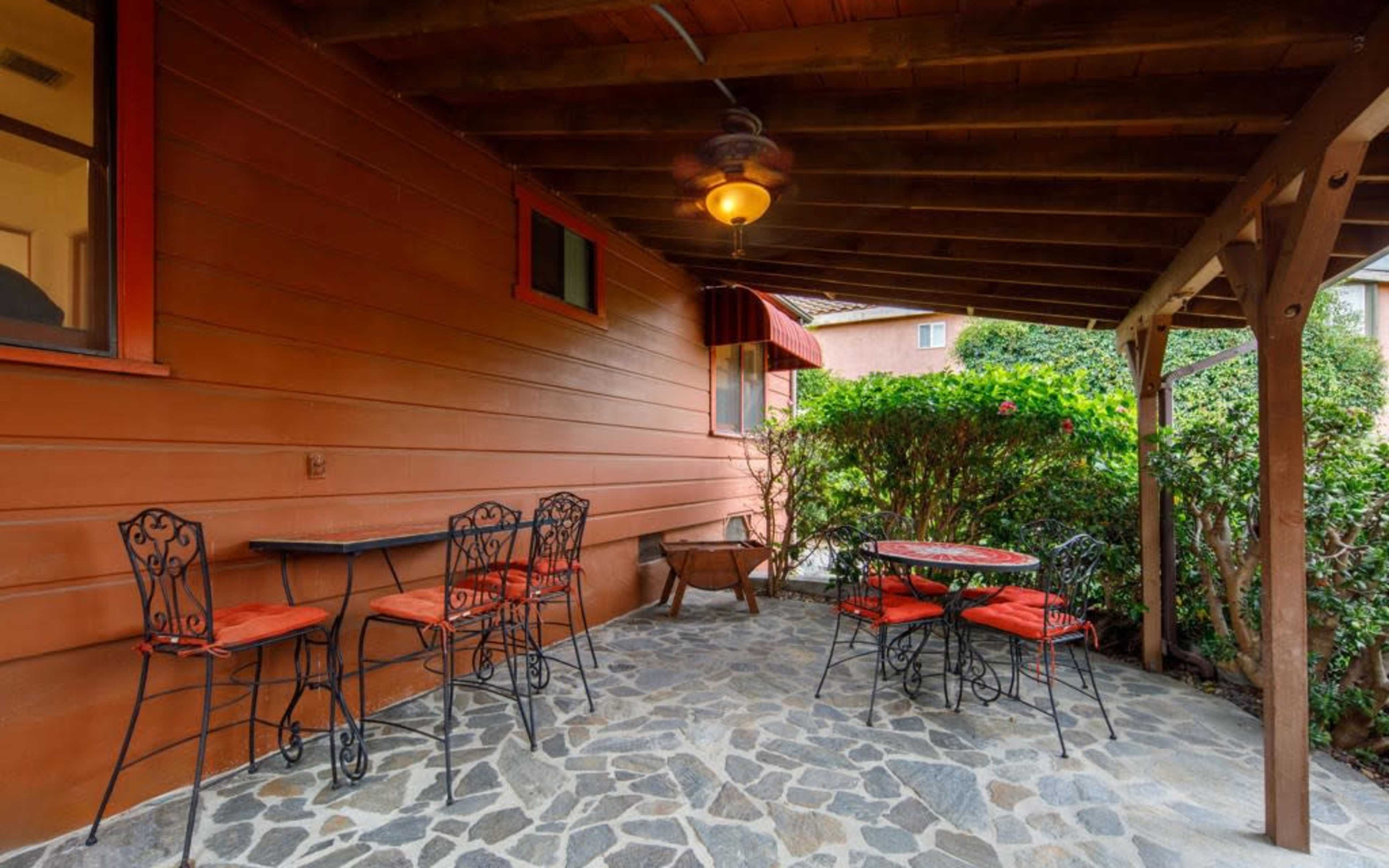 A covered patio with stone flooring, featuring a table and chairs along with a small garden area.
