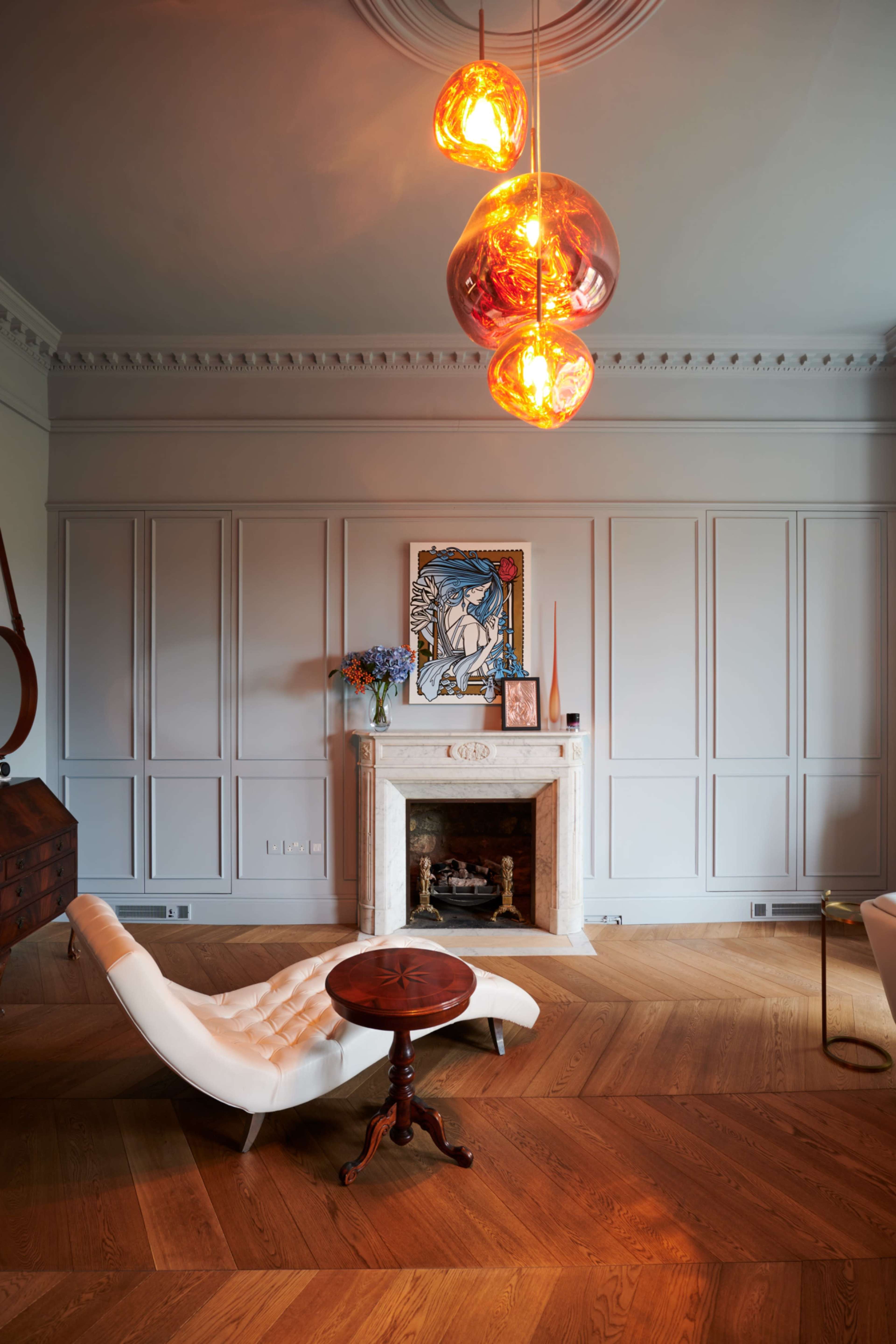 The room features a modern design with a white lounge chair, a small round table, a fireplace with a framed art piece above it, and pendant lights hanging from the ceiling.