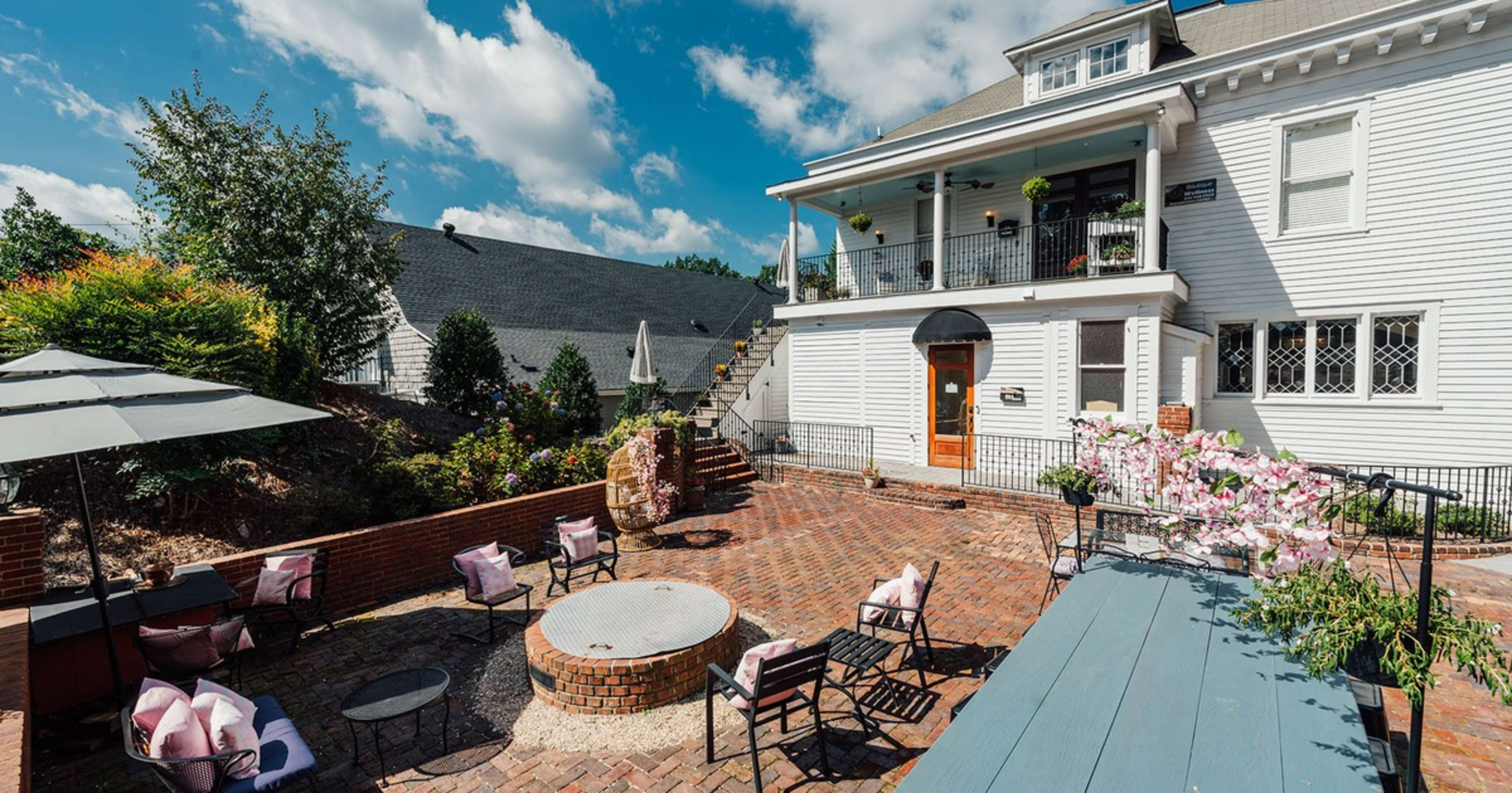 A brick patio with seating and a fire pit is situated outside a large white house, featuring a balcony and landscaped garden.