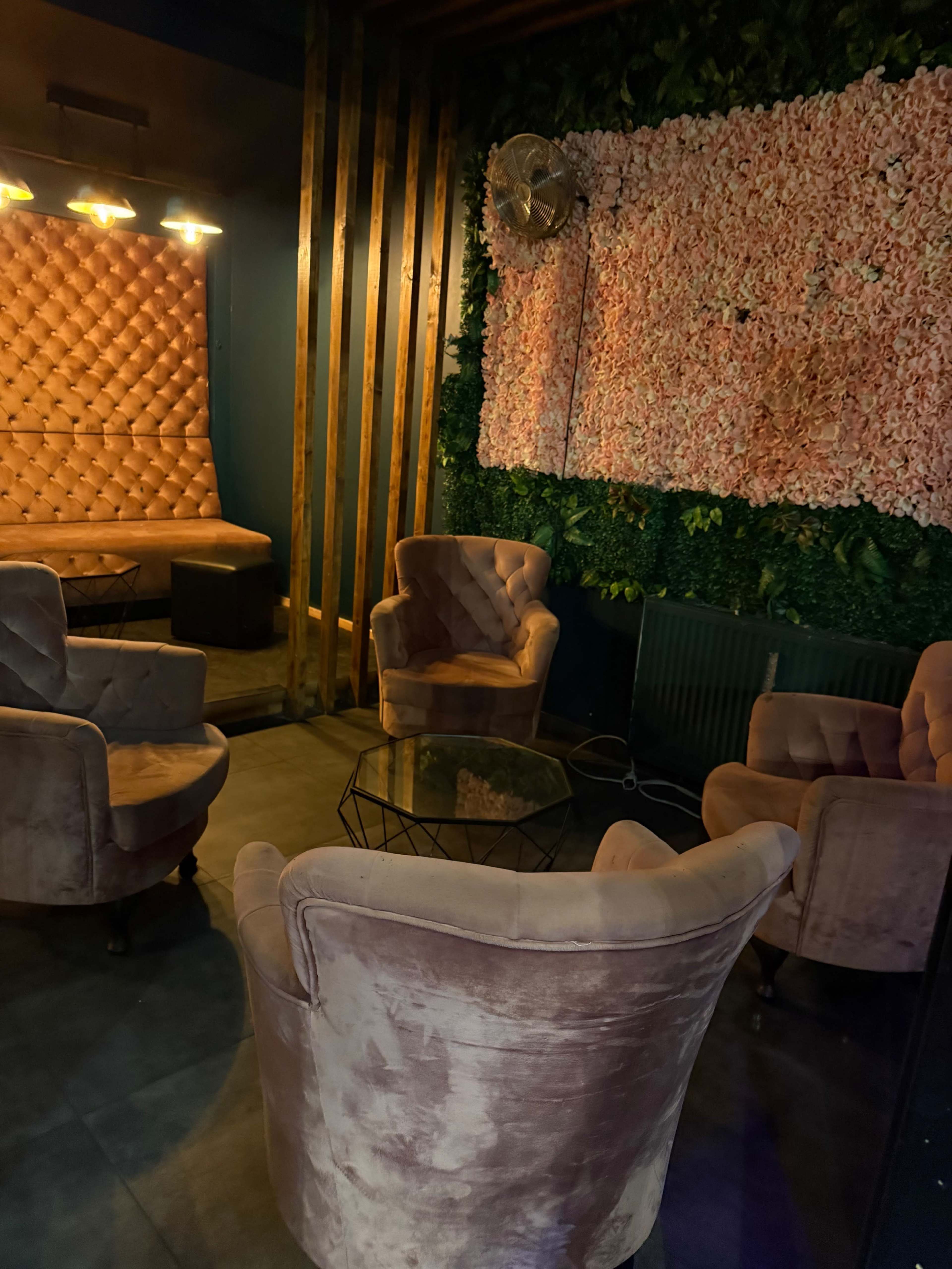 The image shows a cozy lounge area with plush, light-colored chairs arranged around a glass table, set against a backdrop of a floral wall and wooden accents.