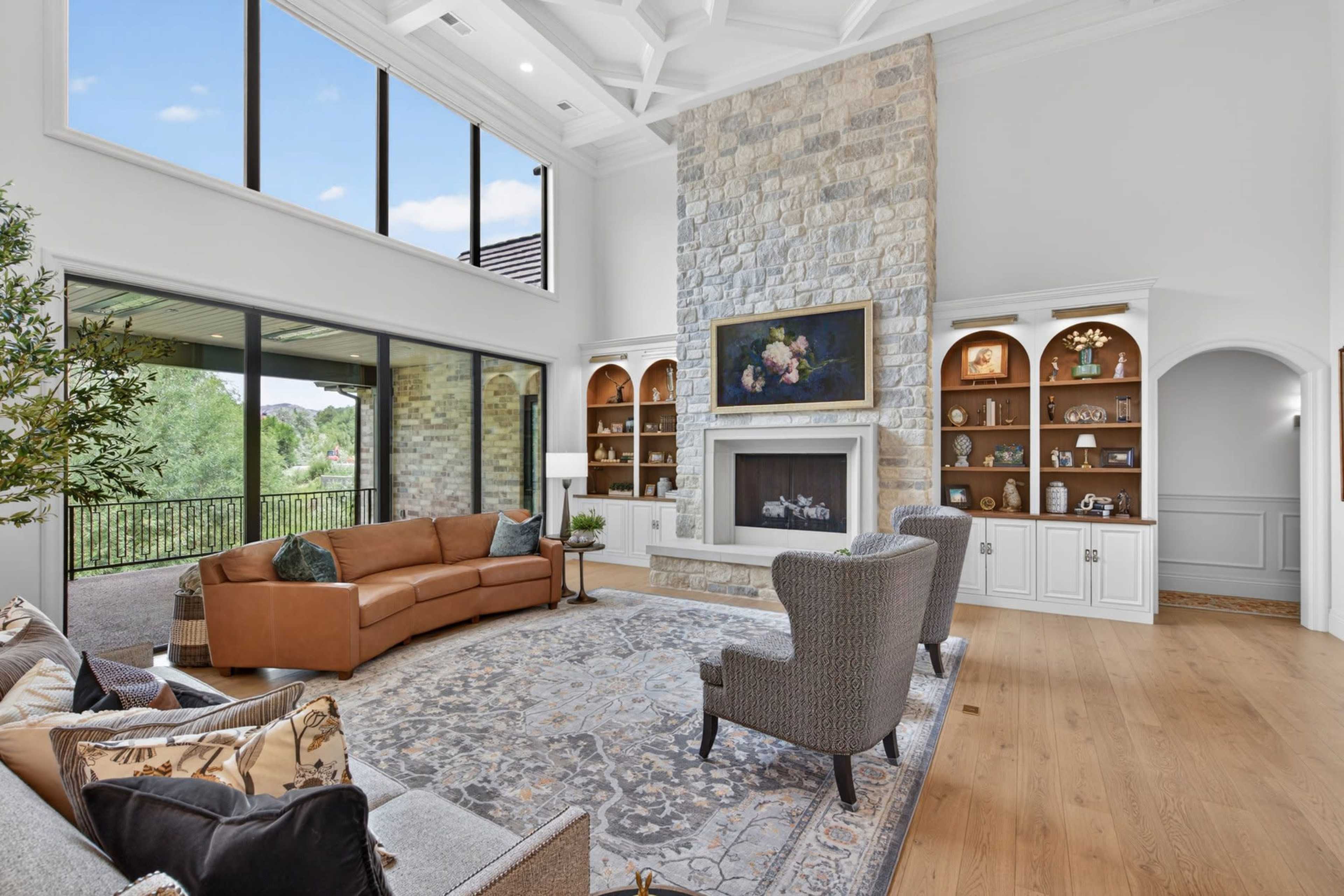 The living room features a stone fireplace, large windows, and a combination of a brown leather sofa and gray armchairs arranged around a decorative area rug.
