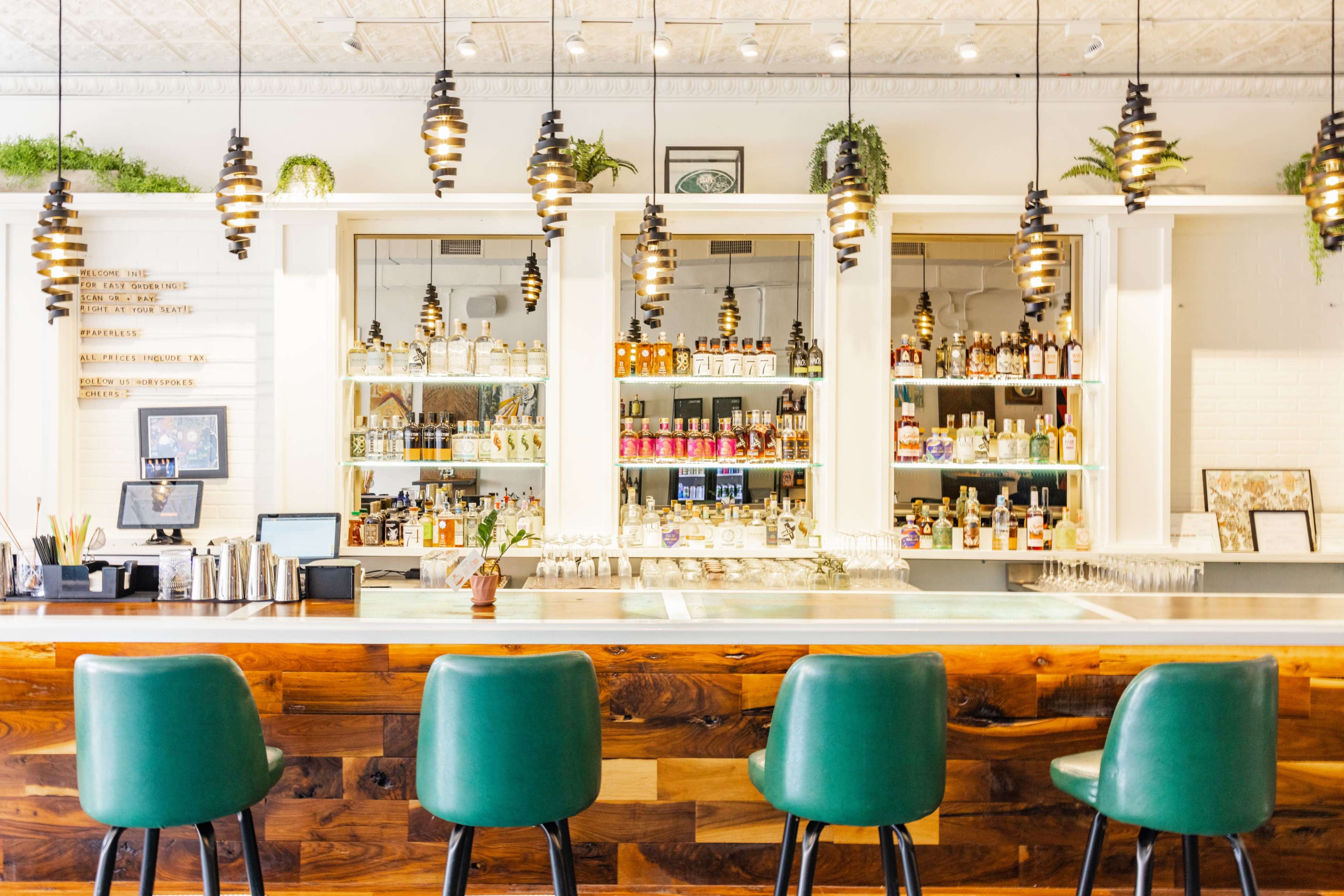 A well-lit bar with a wooden counter, green upholstered stools, and shelves displaying various bottles of alcohol.