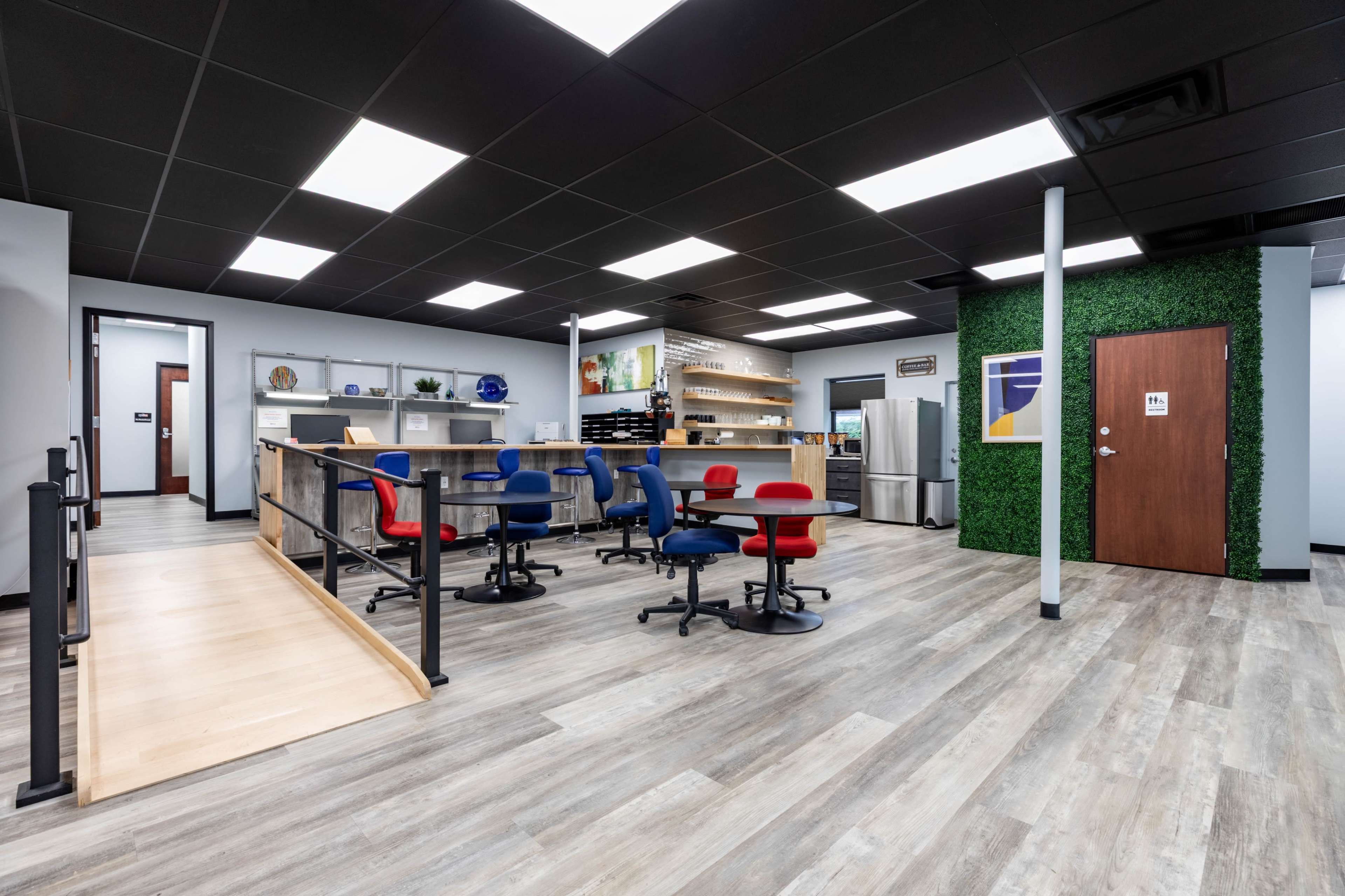 The image shows a modern office space featuring a kitchenette area with a counter, several work stations with blue and red chairs, and green wall accents.