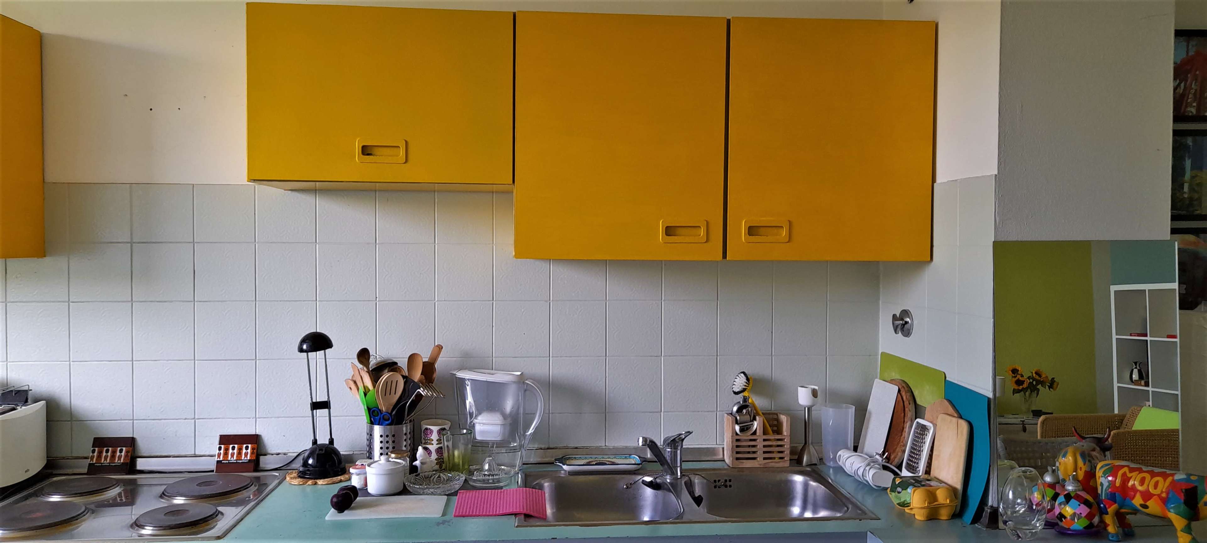 The image shows a kitchen with yellow cabinets above a sink and countertop, featuring cooking utensils and appliances.