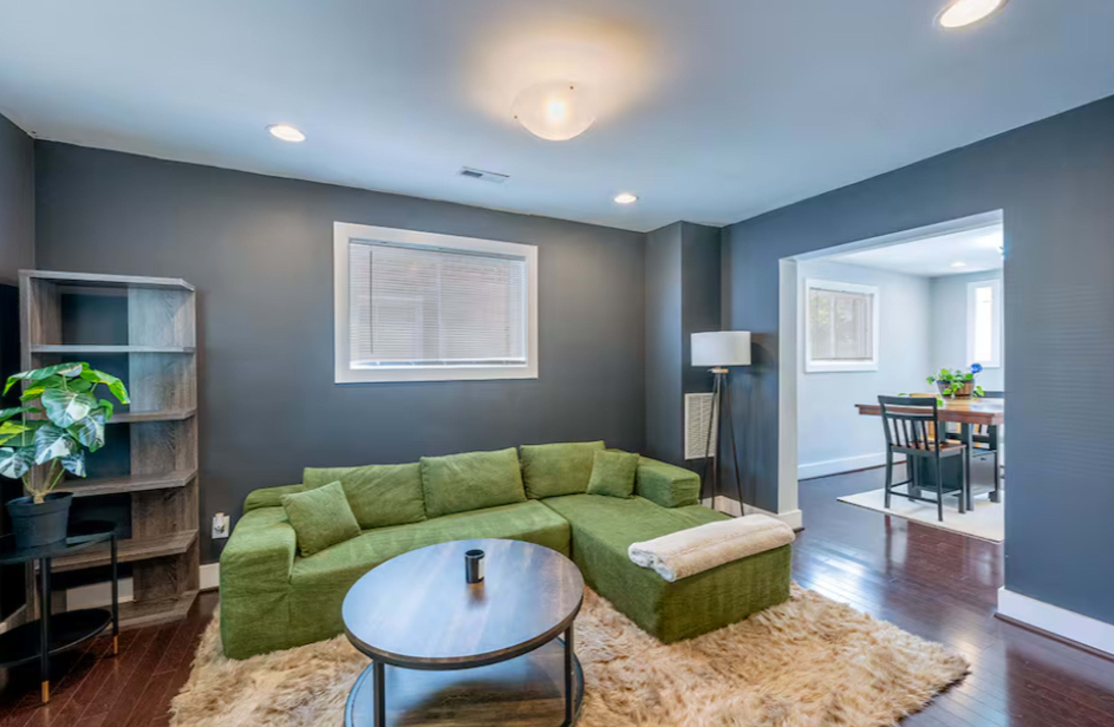 A modern living room features a green sectional sofa, a round coffee table, and a small shelving unit, with a view of a dining area through an open doorway.