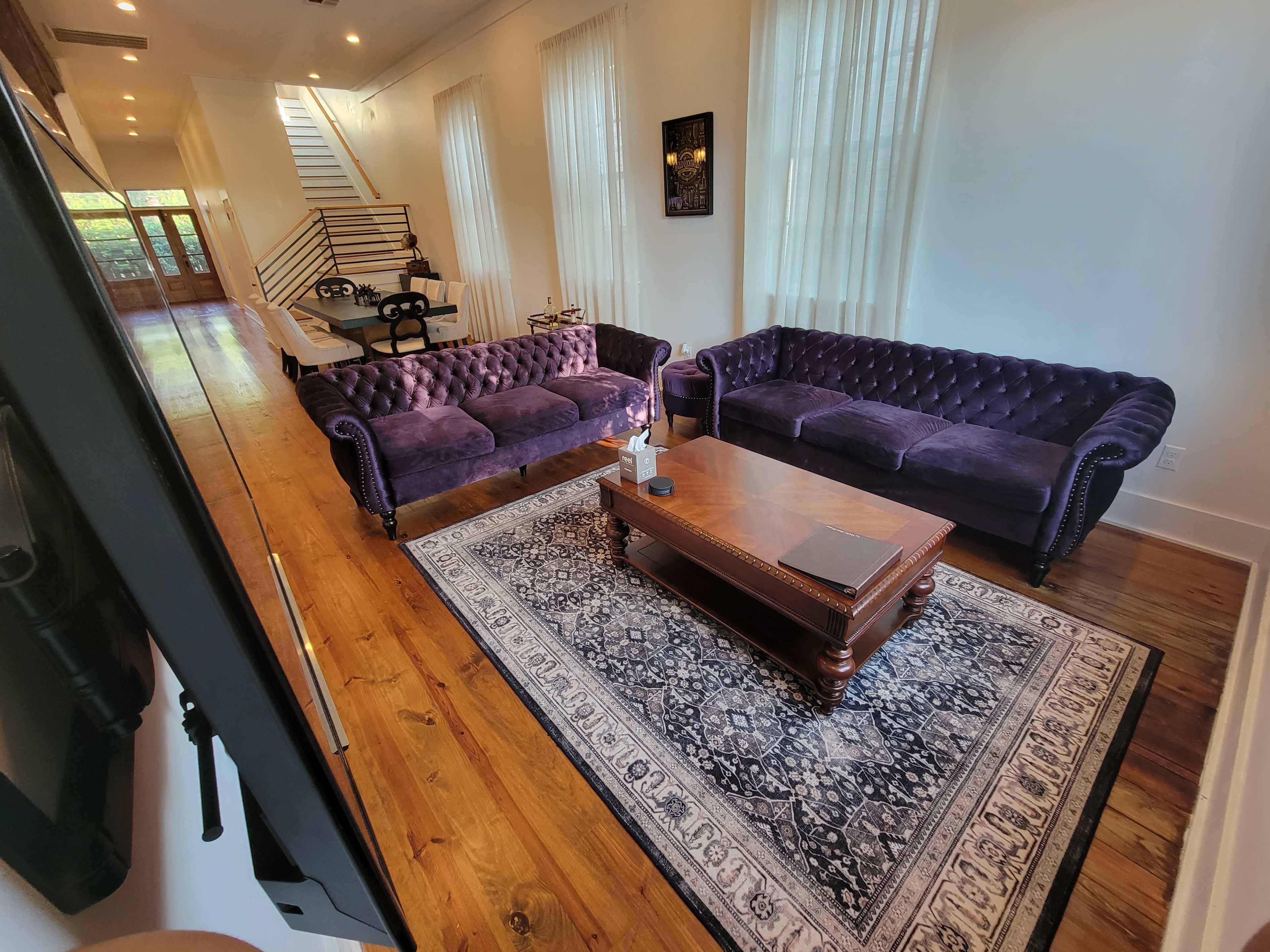 The image shows a living room with two dark purple sofas, a wooden coffee table, and a patterned rug, all set in a bright space with large windows and wooden flooring.