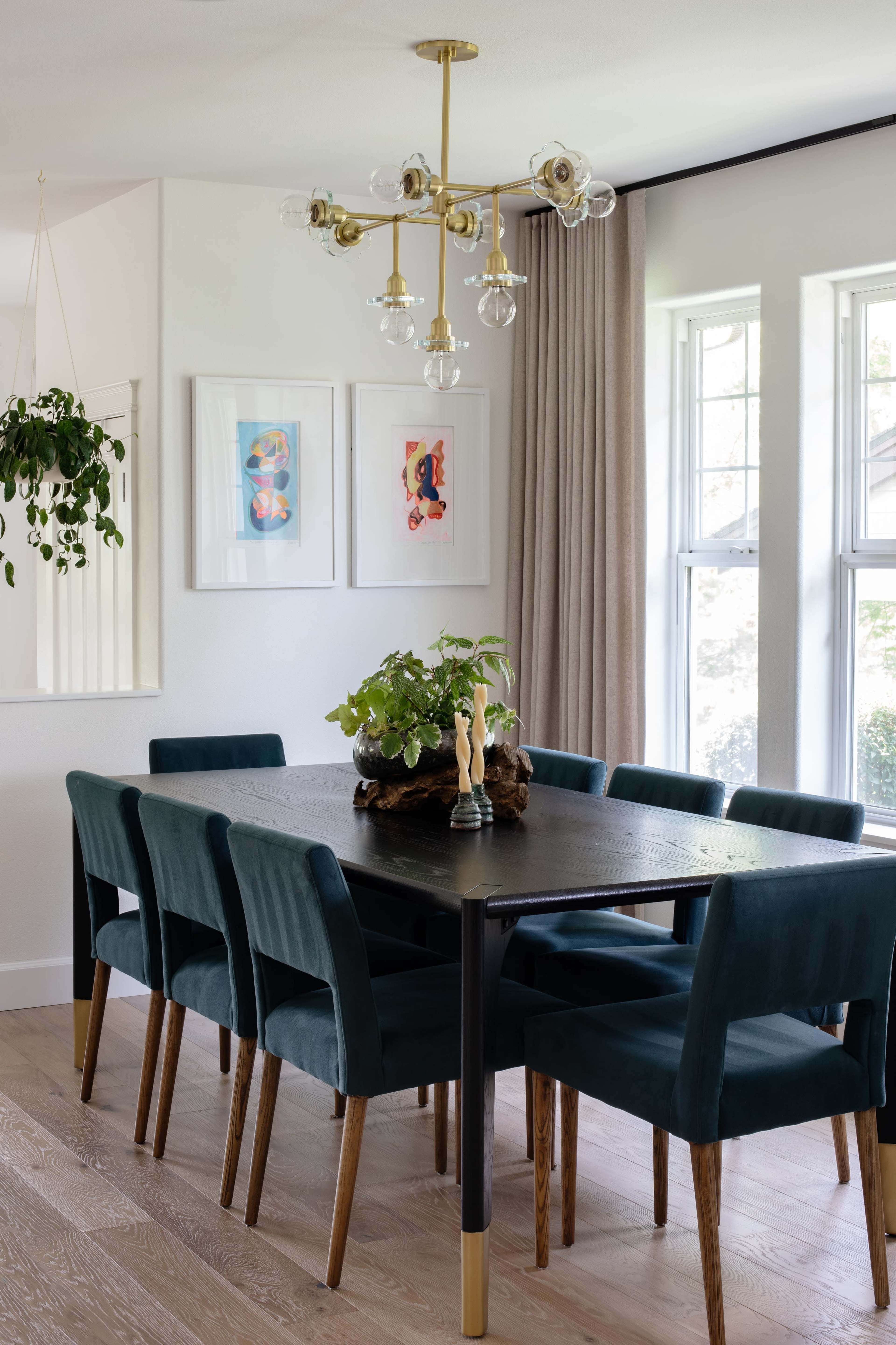 A modern dining room features a black table surrounded by green upholstered chairs, a decorative centerpiece, and large windows providing natural light.