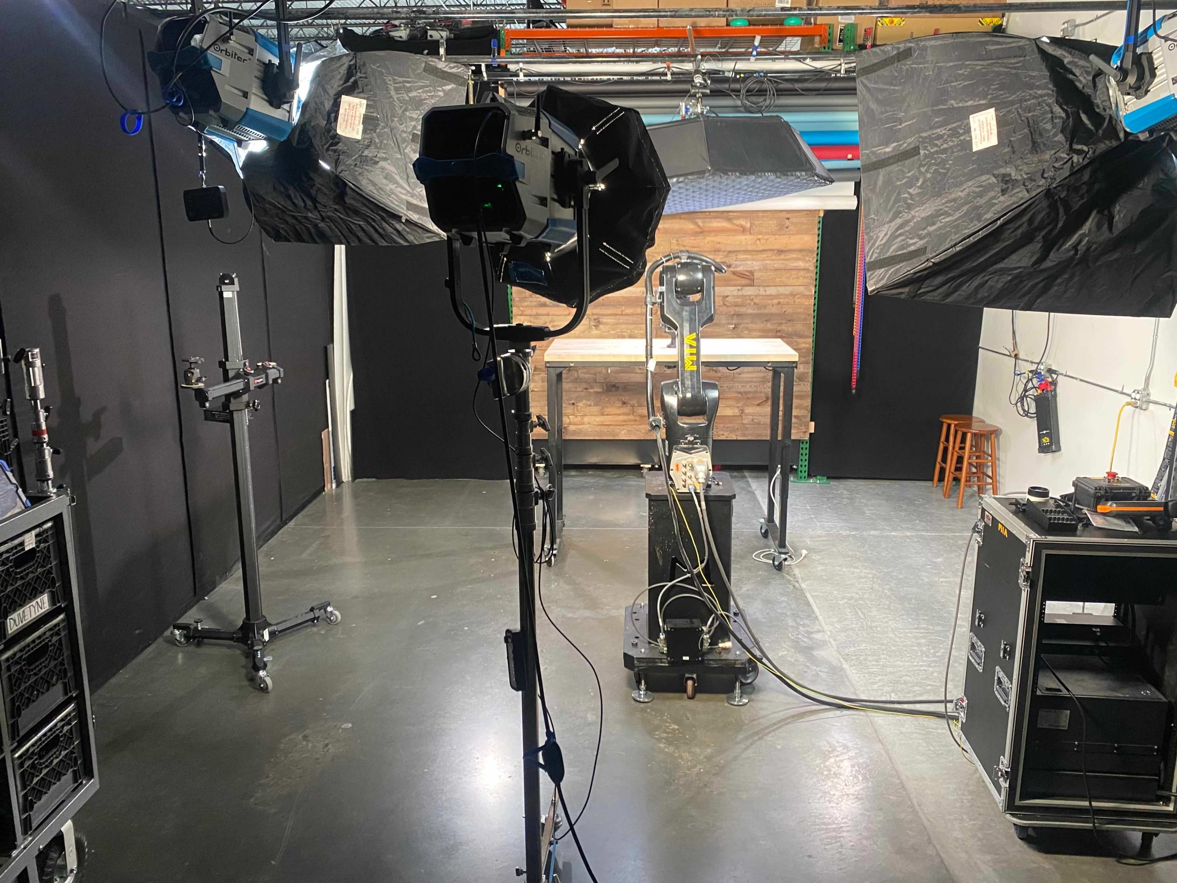A studio setup with two large softbox lights, a robotic arm in the center, and various equipment arranged around a wooden tabletop in a black-walled space.