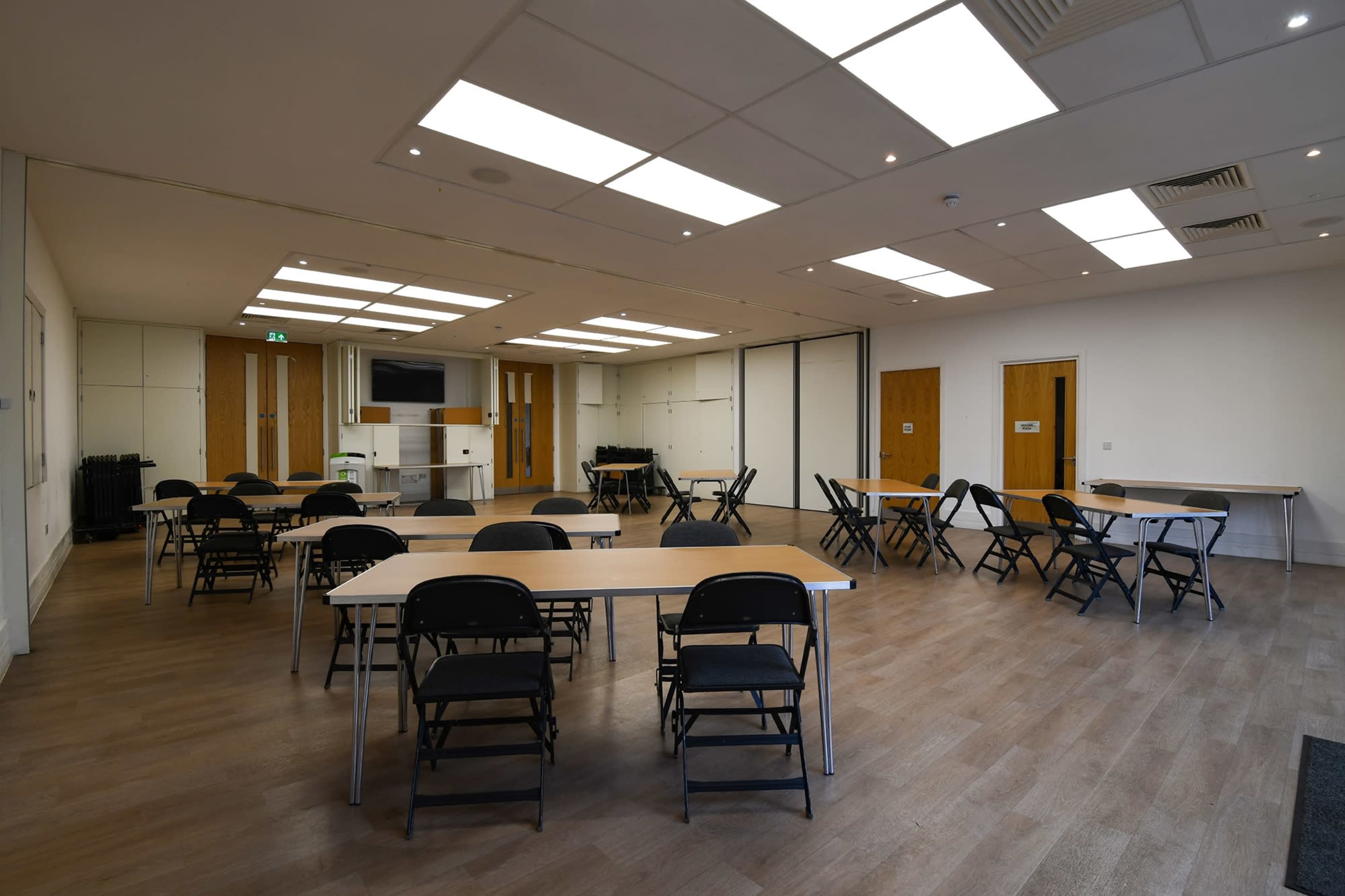 A spacious meeting room features several tables and black chairs arranged throughout the area, with large windows and bright overhead lights.