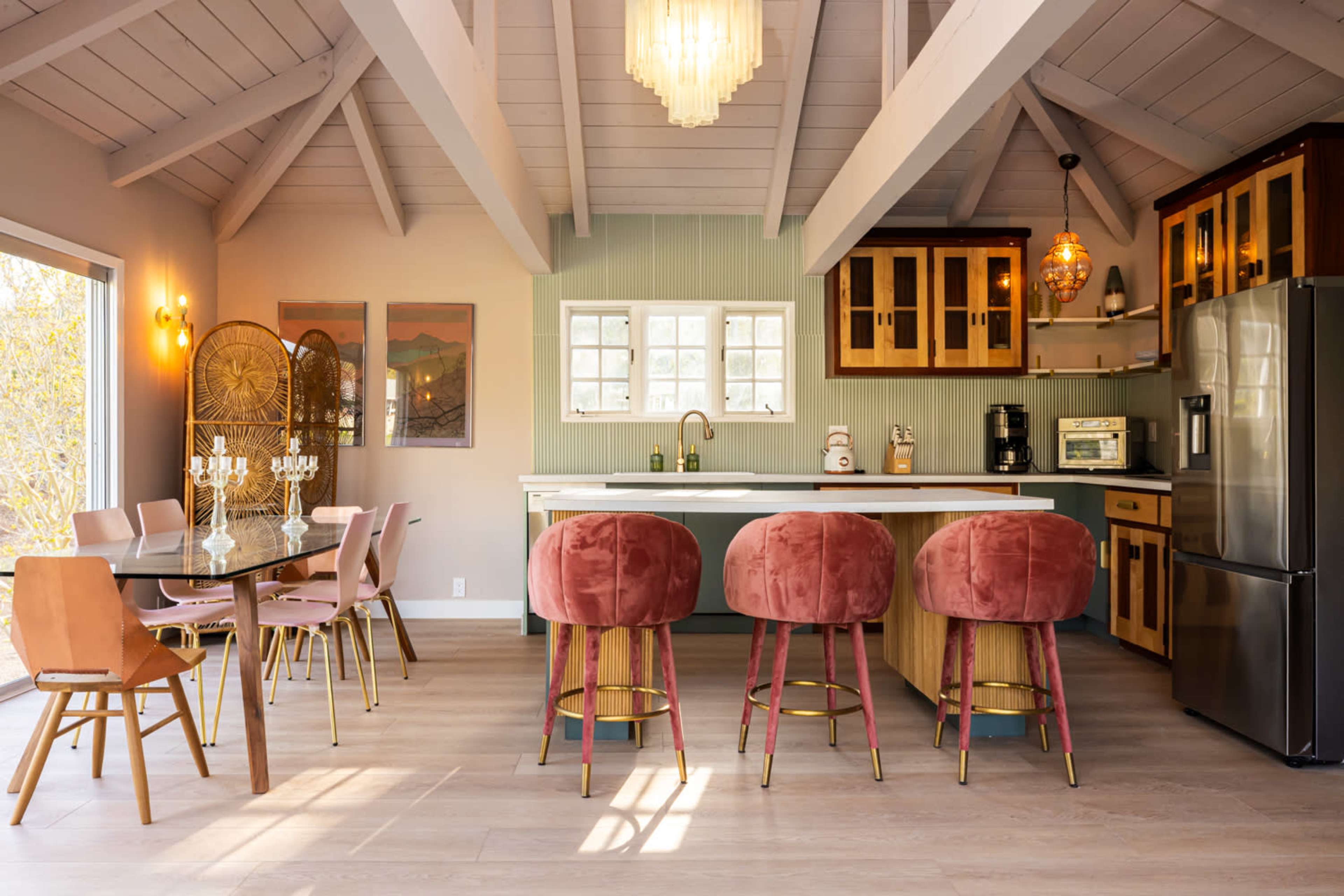The image shows a modern kitchen and dining area featuring a large island with bar stools, a dining table, and large windows that allow natural light to fill the space.