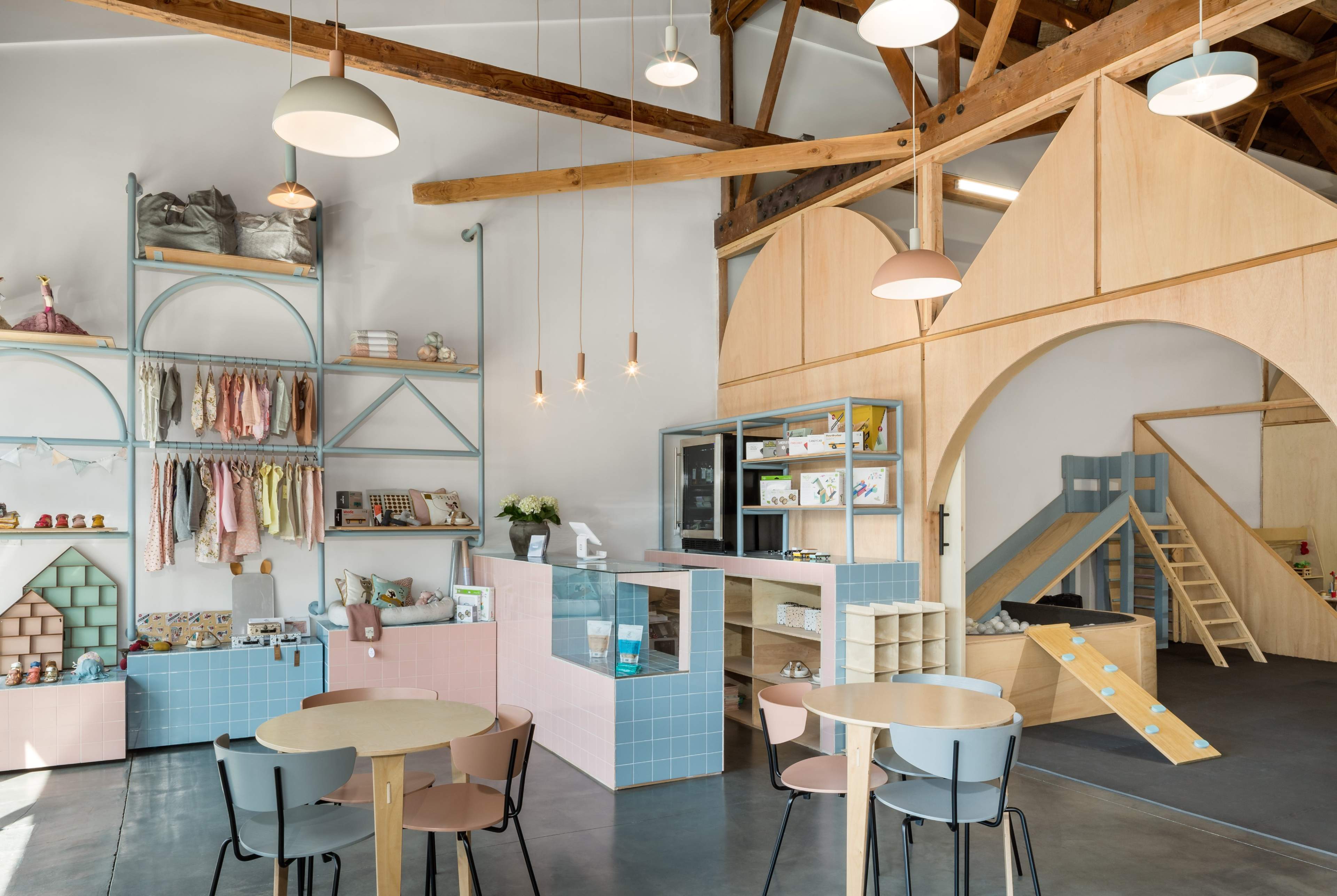 A modern children's store featuring wooden beams, colorful shelving displaying clothes and toys, and a play area with a slide and climbing structure.