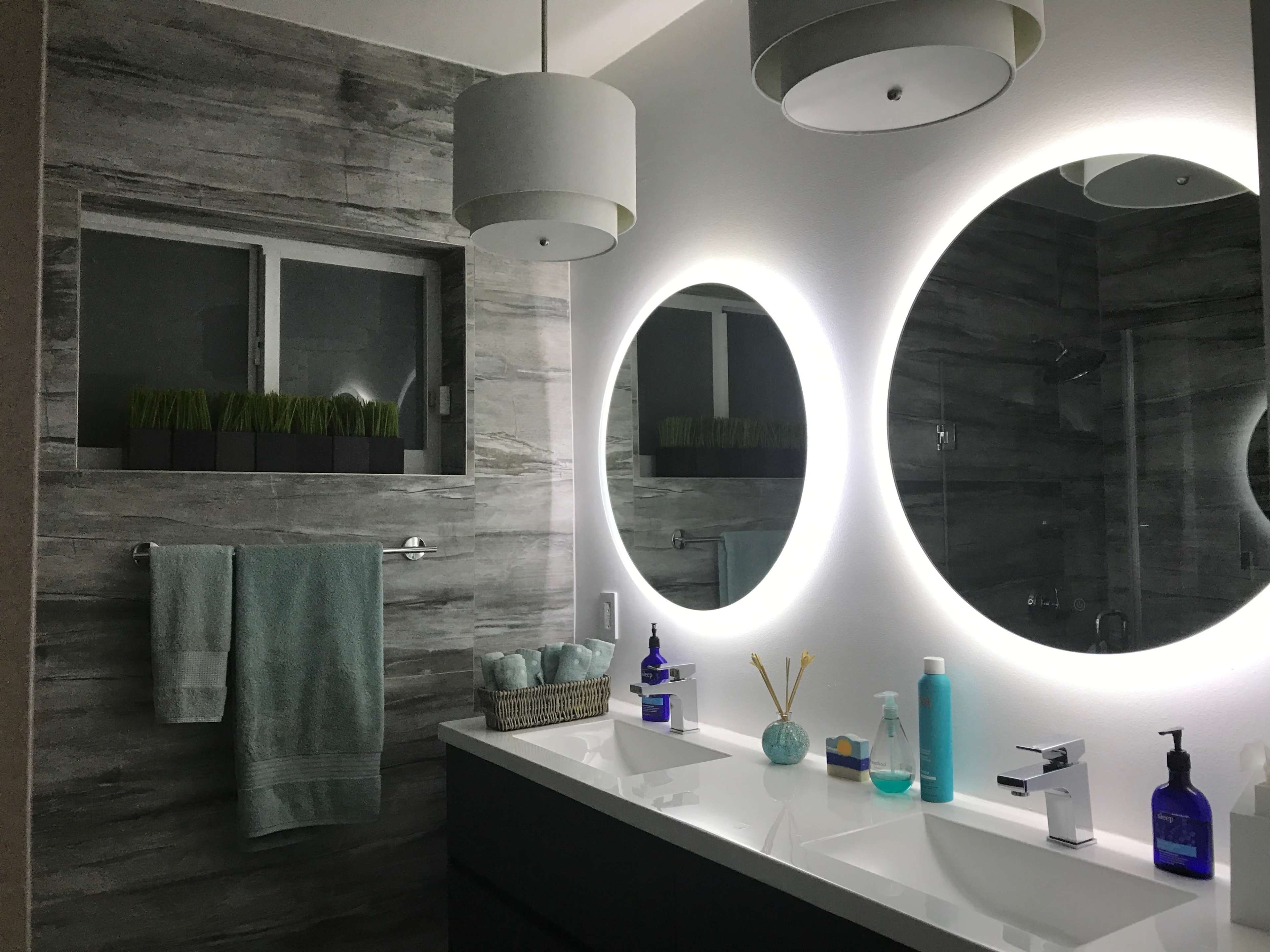 A modern bathroom featuring two circular mirrors with backlighting, a double sink, and decorative plants in a window.