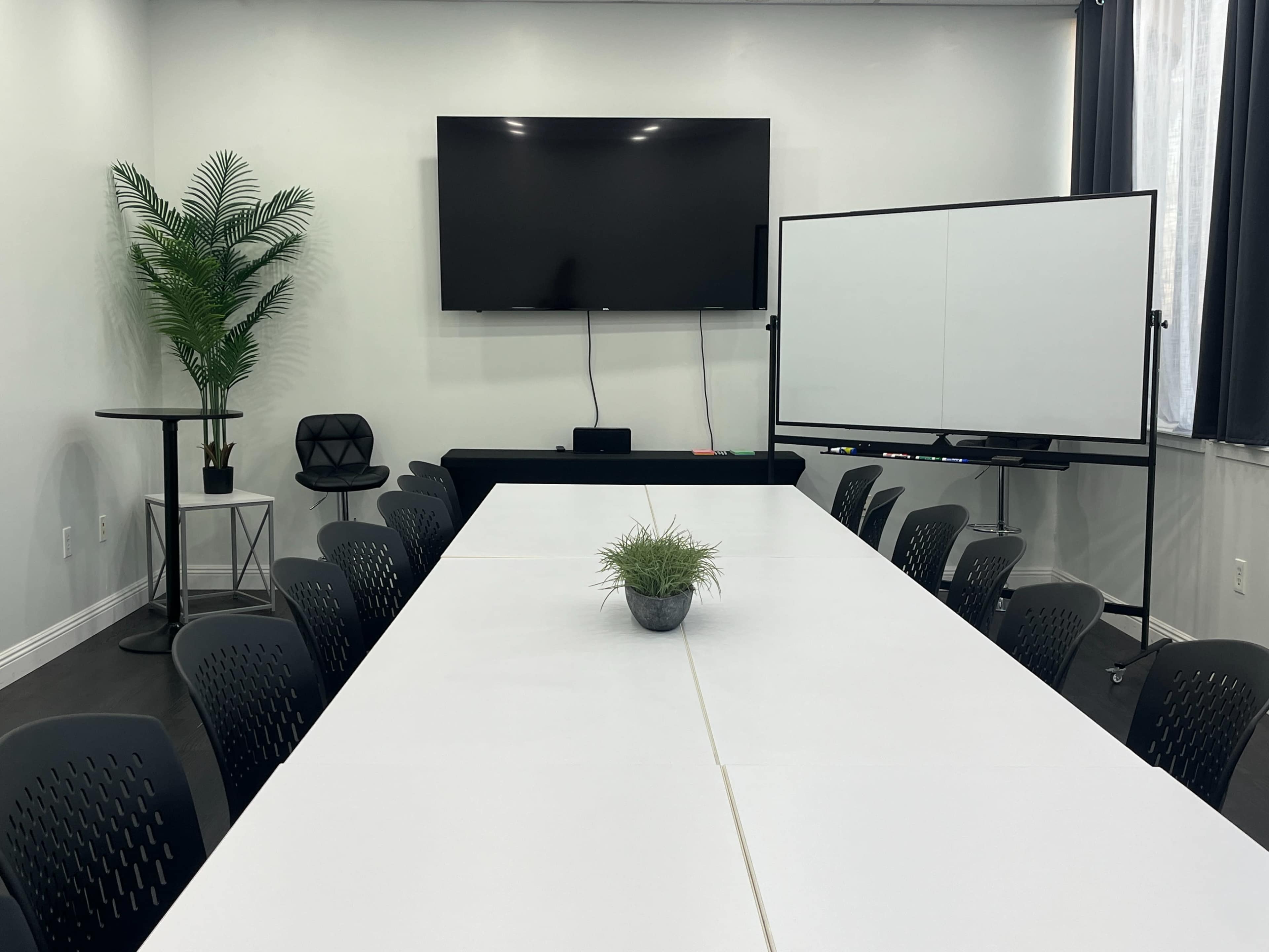 A modern conference room features a large white table surrounded by black chairs, a flat-screen TV on the wall, and a mobile whiteboard.