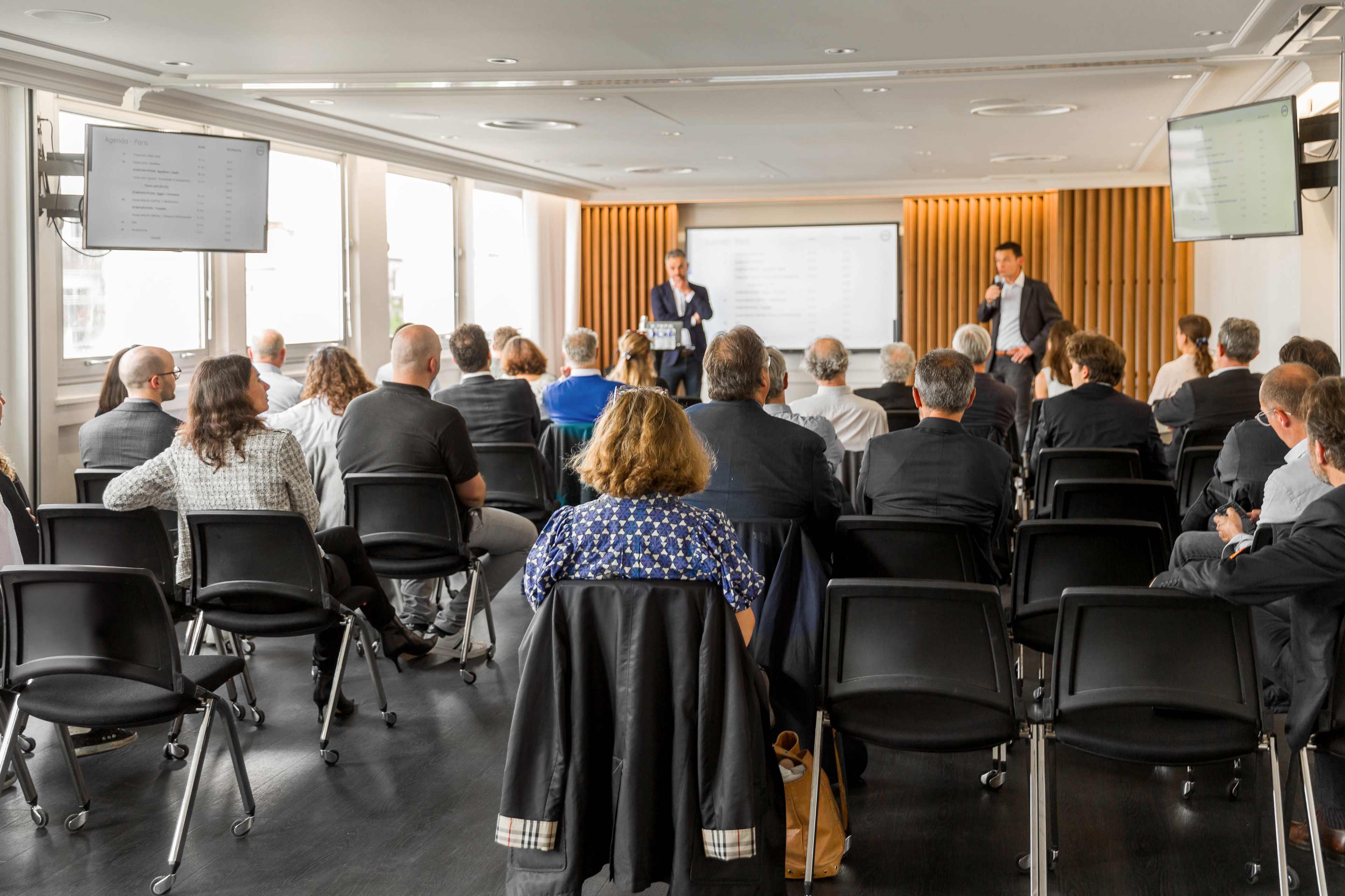 A group of people sits in an auditorium-style setting, facing a speaker presenting at the front of the room.