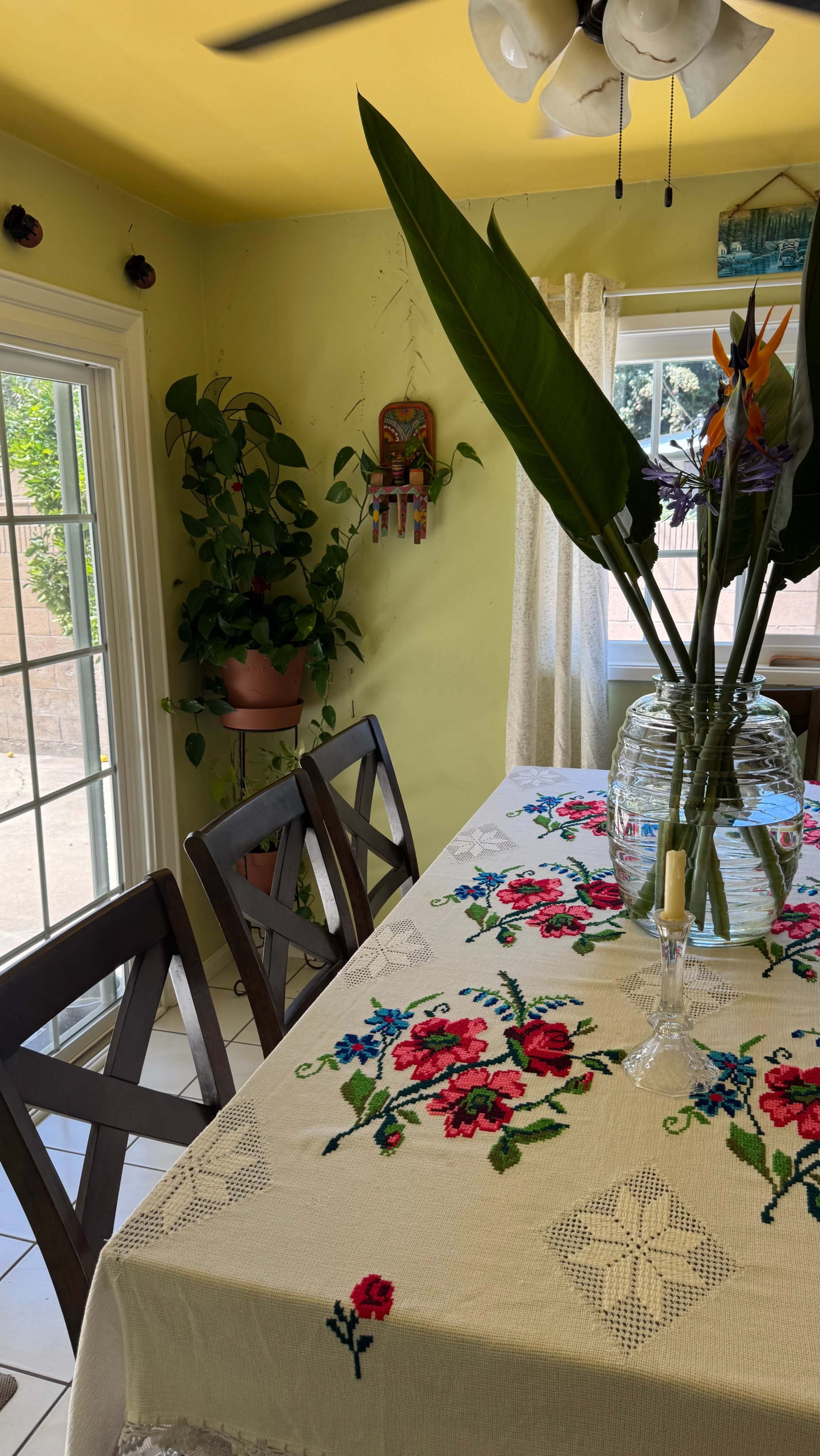 A dining area features a floral tablecloth on a table, two wooden chairs, a large glass vase with tropical flowers, and a potted plant by a bright yellow wall.