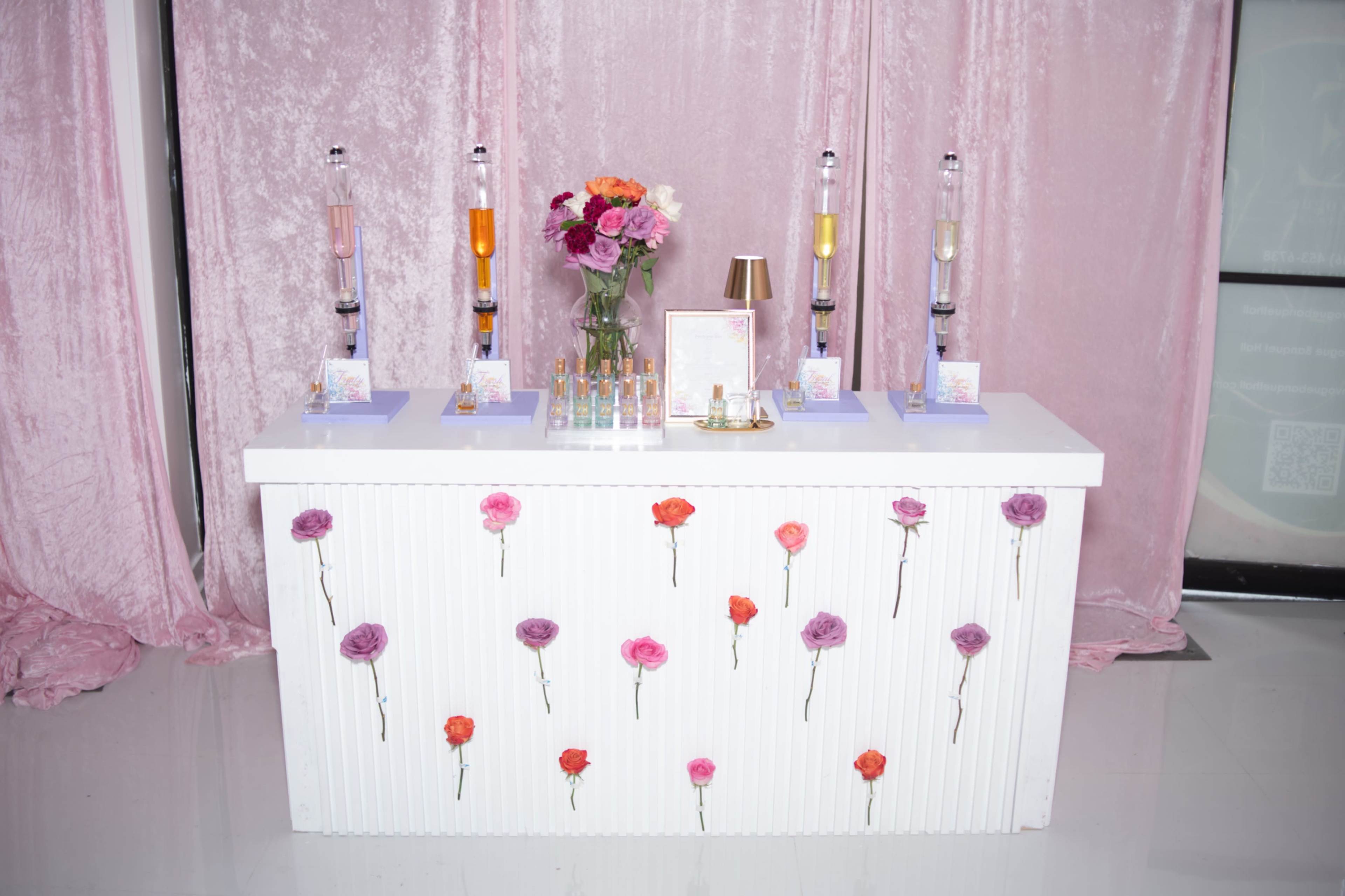 A white table with floral decorations and several glass containers filled with colorful liquids stands in front of a pink backdrop.
