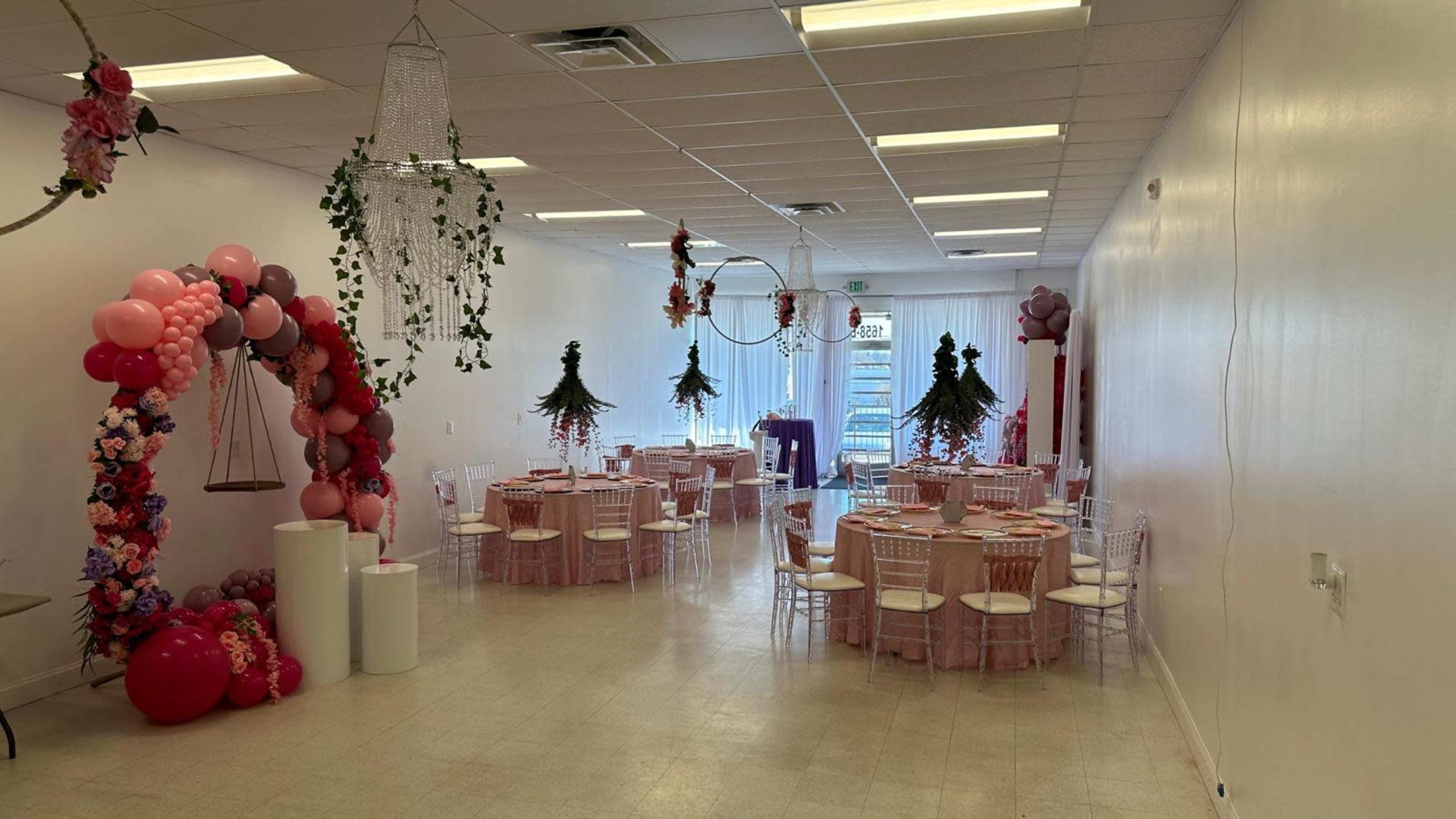 The image shows a brightly lit event space set up for a celebration, featuring round tables with pink tablecloths, decorated with floral arrangements and balloons, with an entry arch made of balloons and flowers.