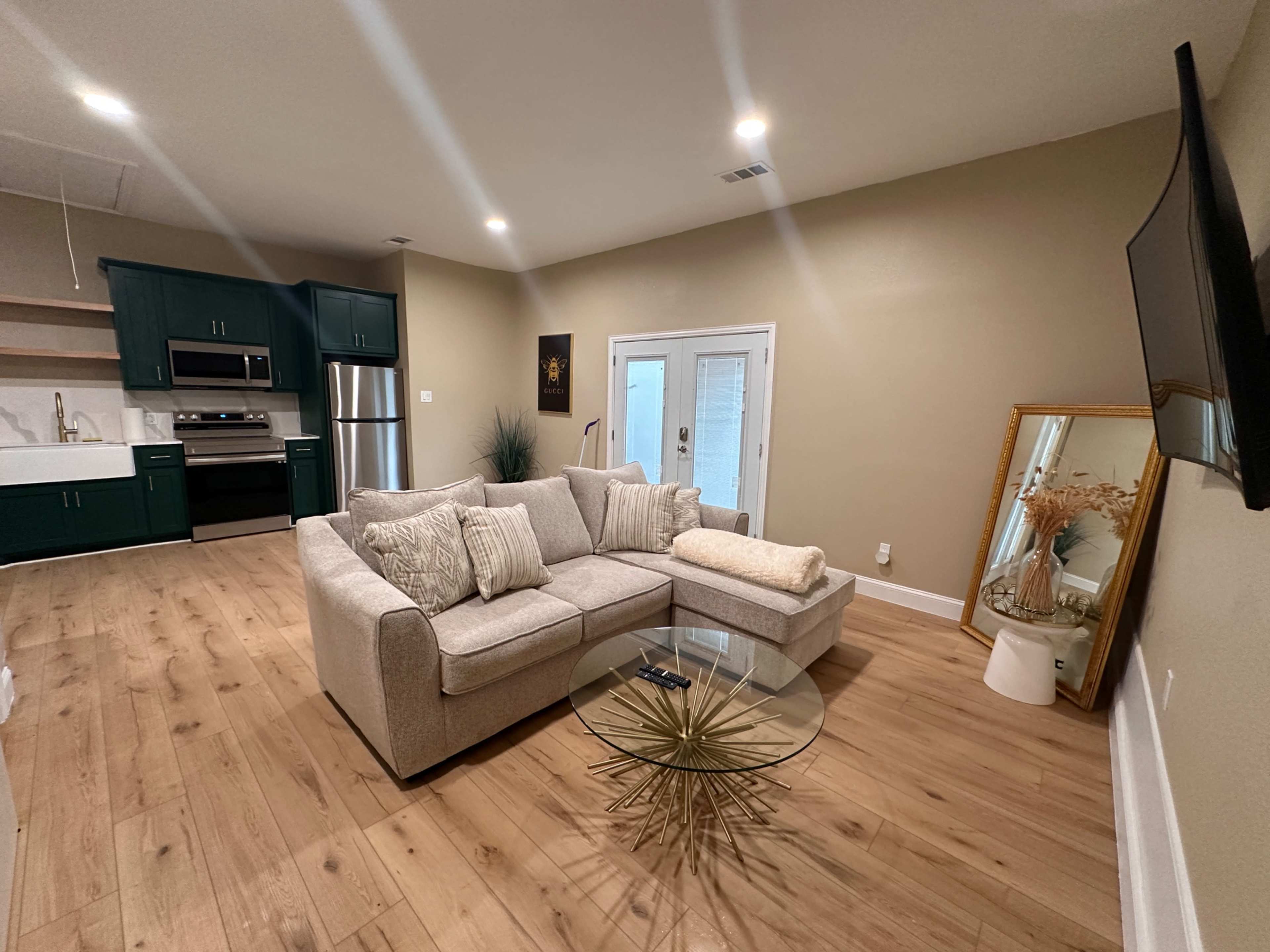 The living space features a light-colored sectional sofa, a glass coffee table with a gold starburst base, and a kitchen area with dark green cabinetry.