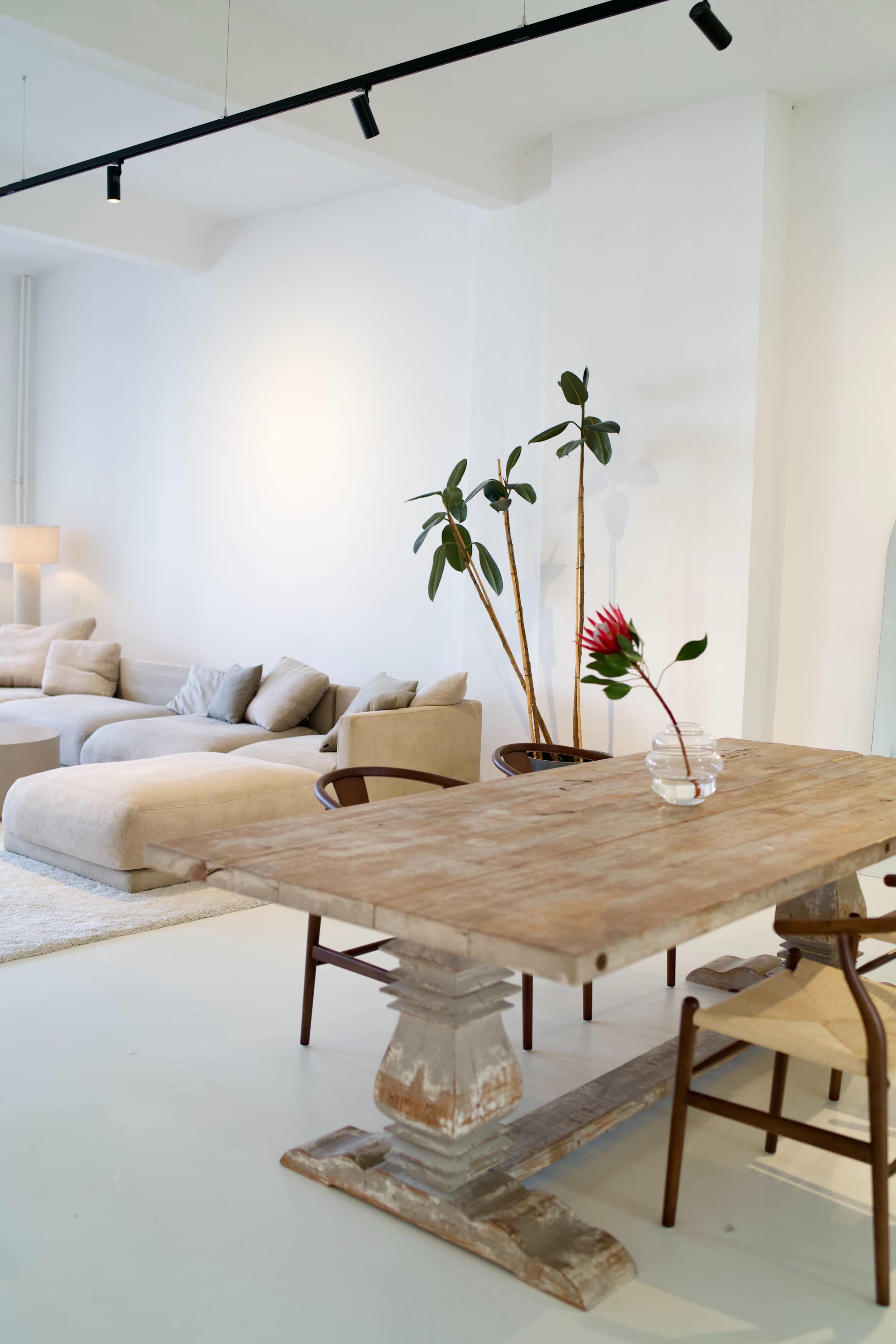 A spacious room features a wooden dining table with chairs, a large beige sectional sofa, and potted plants against a light wall.