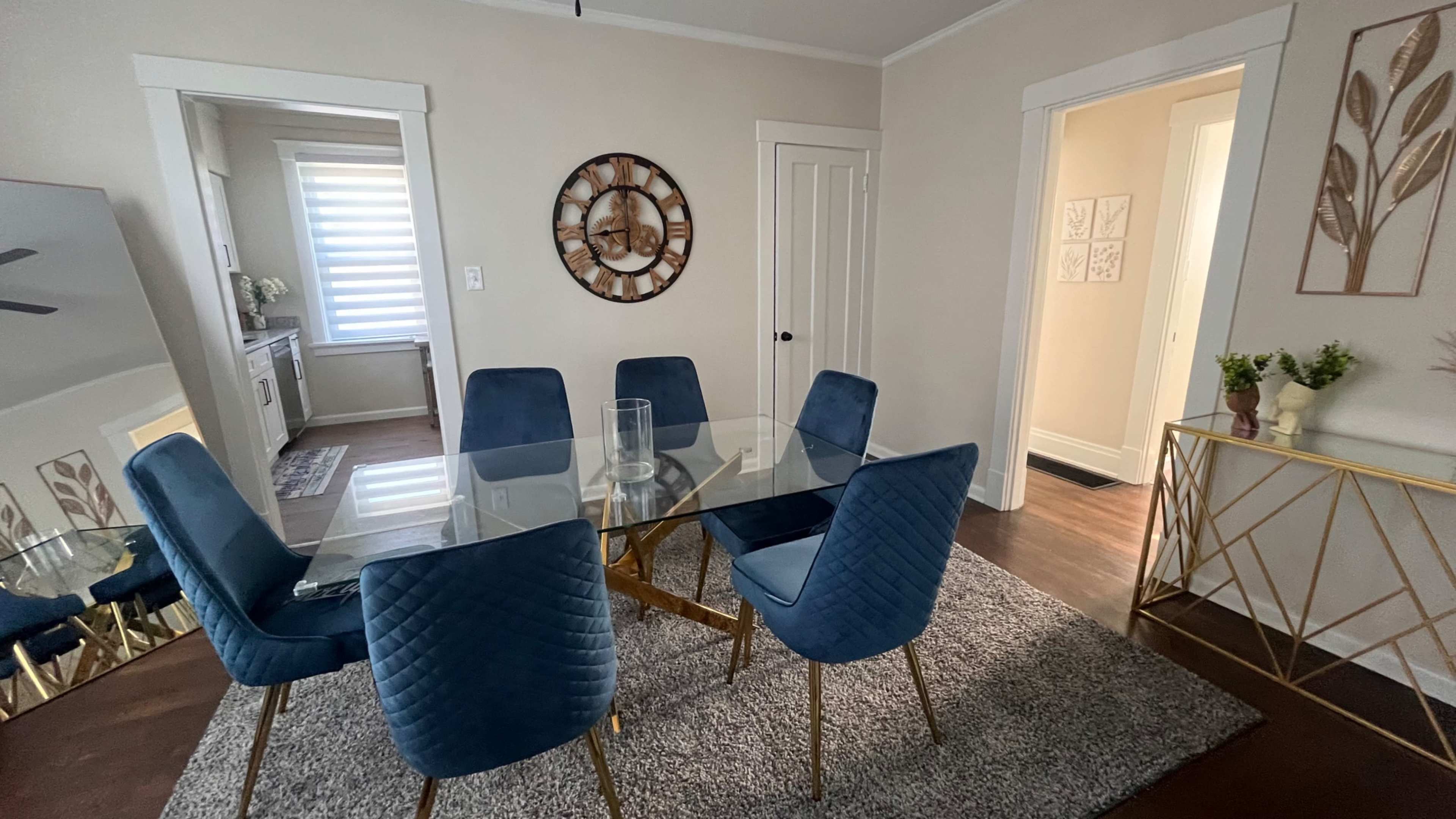 The image shows a dining area with a glass-top table surrounded by six blue chairs on a gray rug, with a wall clock and two doorways visible in the background.