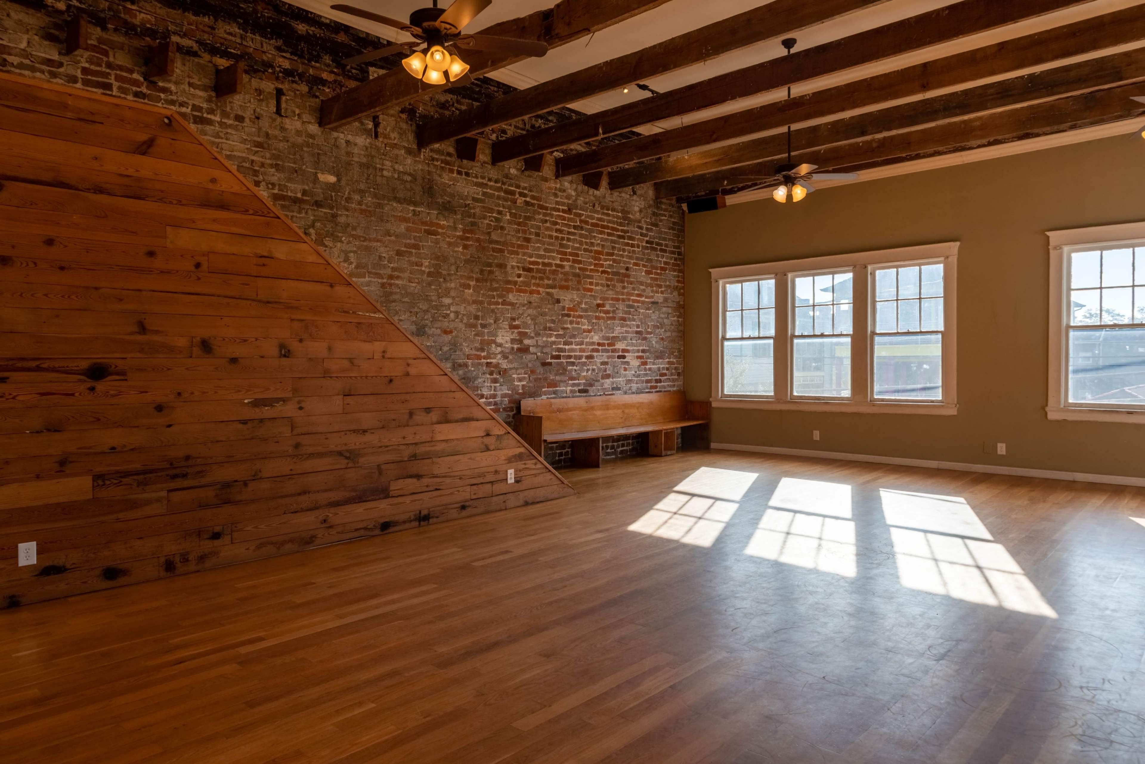 The image shows a spacious, empty room with exposed brick walls, wooden beams on the ceiling, and large windows allowing natural light to enter.