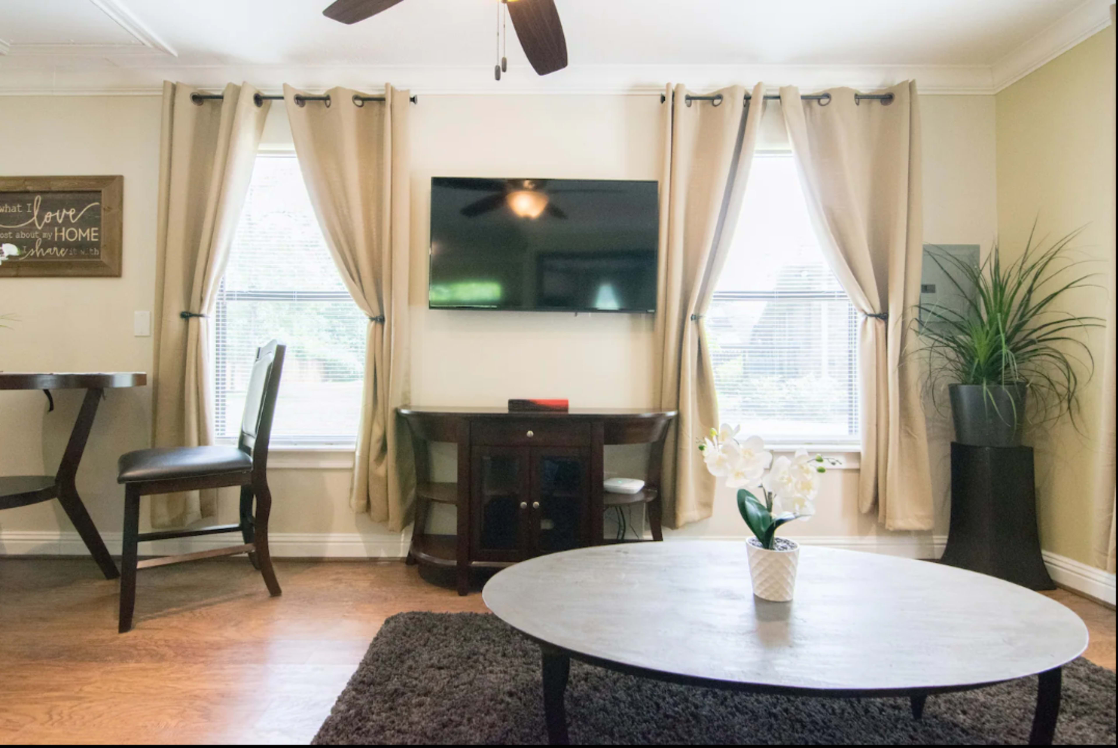 A living room with a television mounted on the wall, a round coffee table, a plant, and two windows with beige curtains.