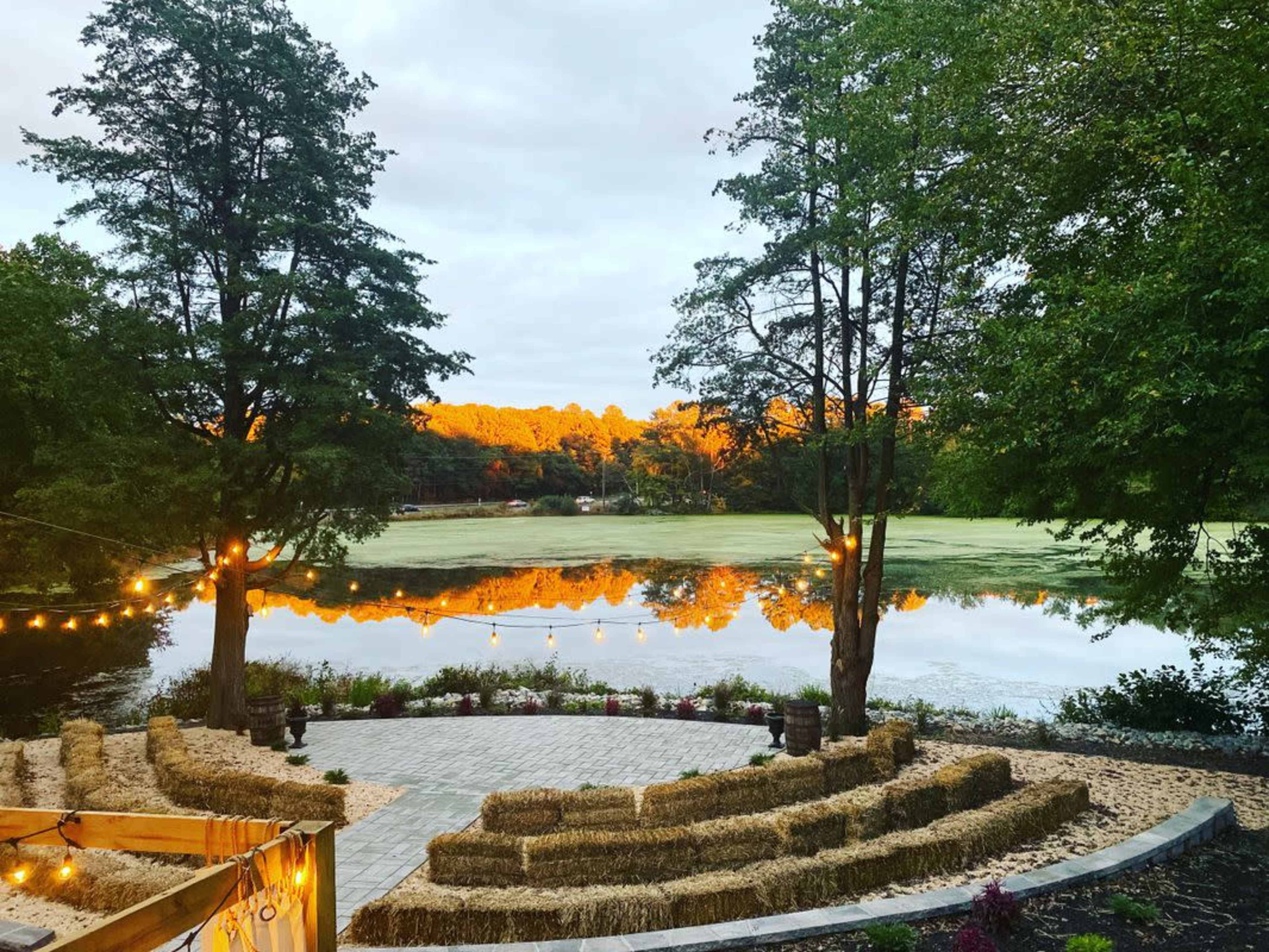 A circular seating area made of hay bales overlooks a tranquil lake bordered by trees, reflecting the orange tones of the sunset.