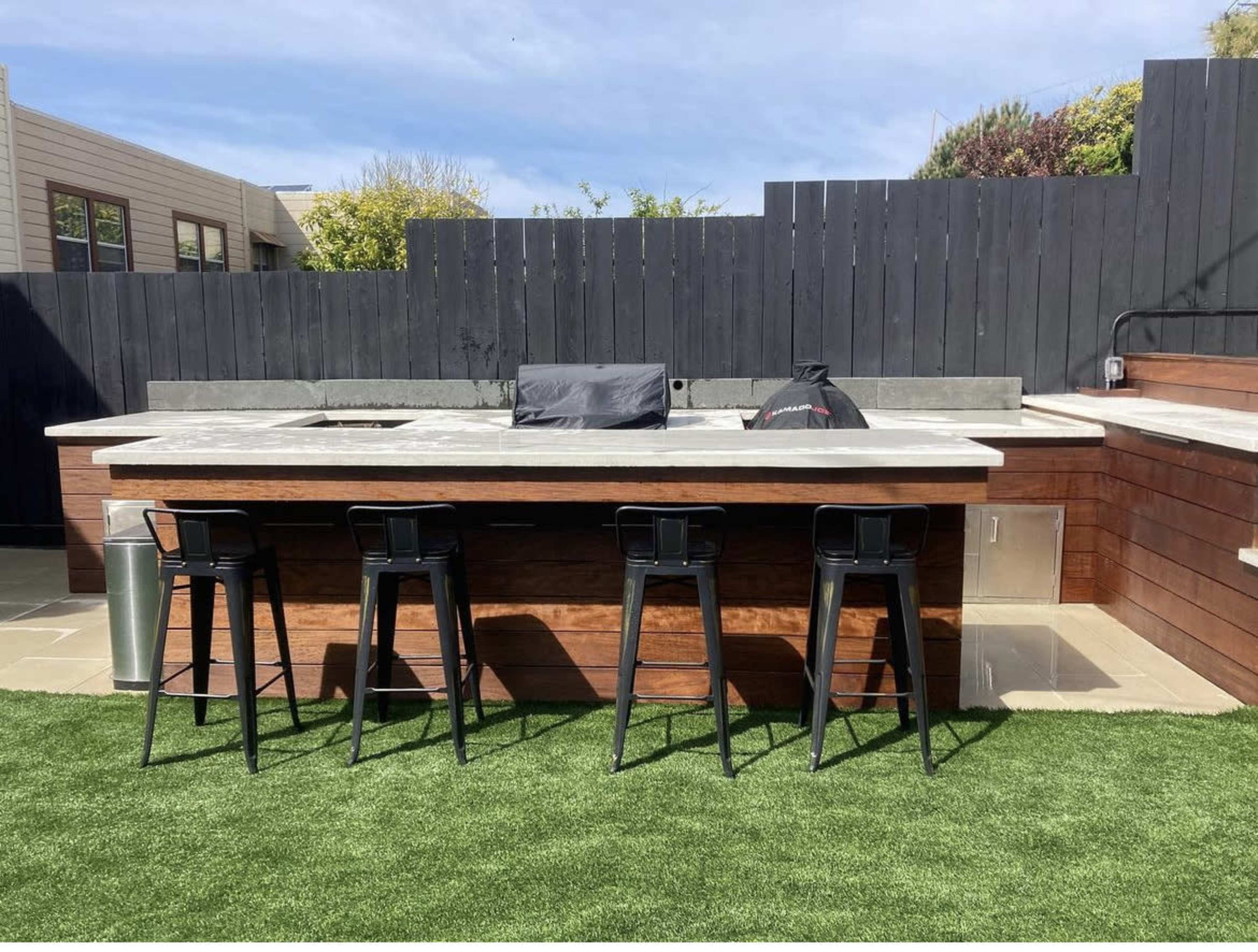 A modern outdoor bar with a concrete countertop, stainless steel appliances, and four black stools on a grassy area.