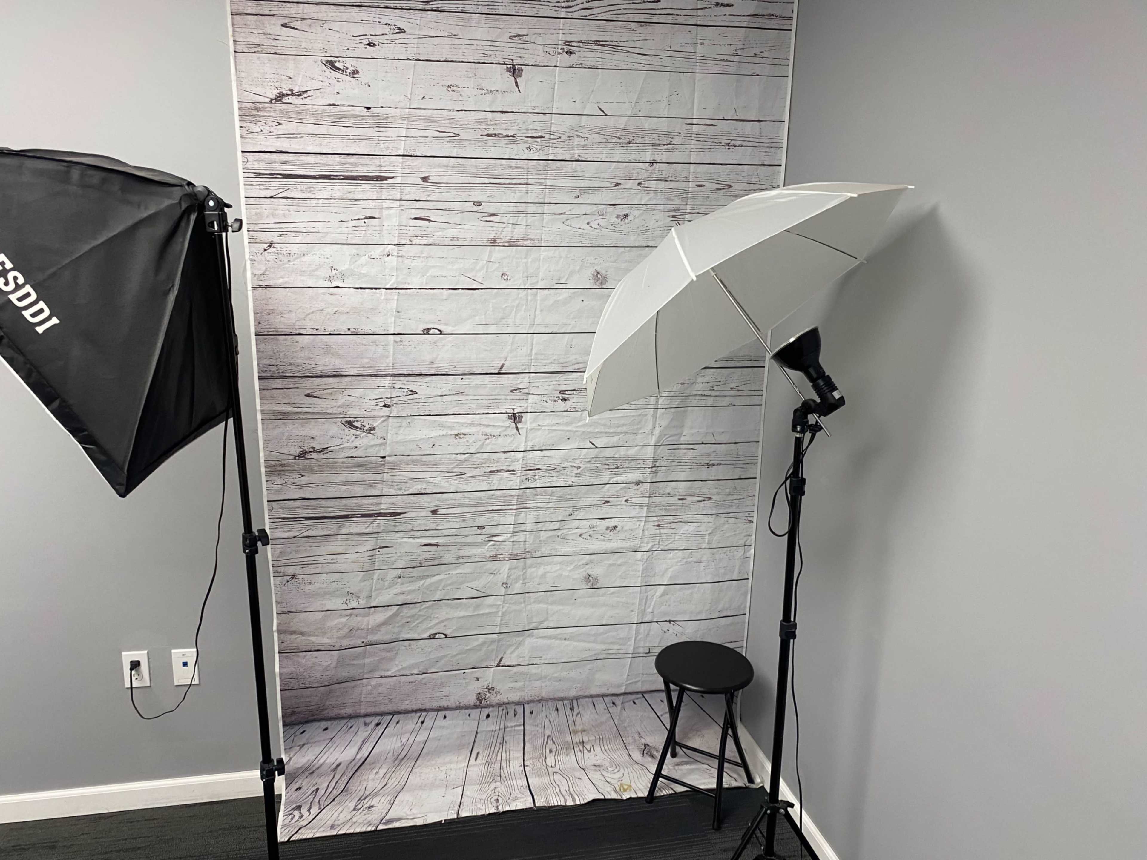 The image shows a photography setup featuring a backdrop of wooden planks, a lighting umbrella, and a black stool.