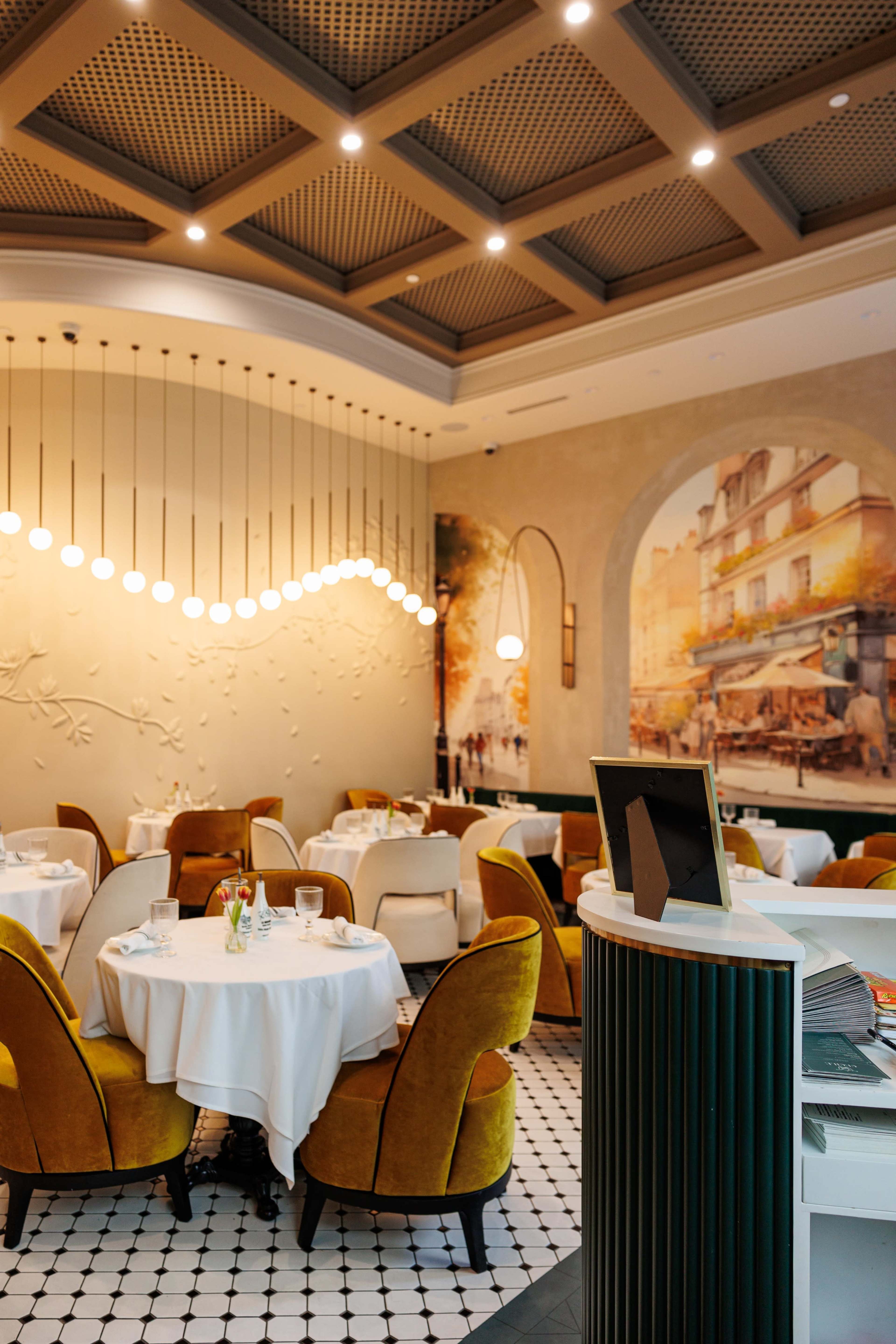 The image shows an elegant restaurant interior with round tables covered in white tablecloths, surrounded by yellow upholstered chairs, and a mural on the wall depicting a Parisian scene.