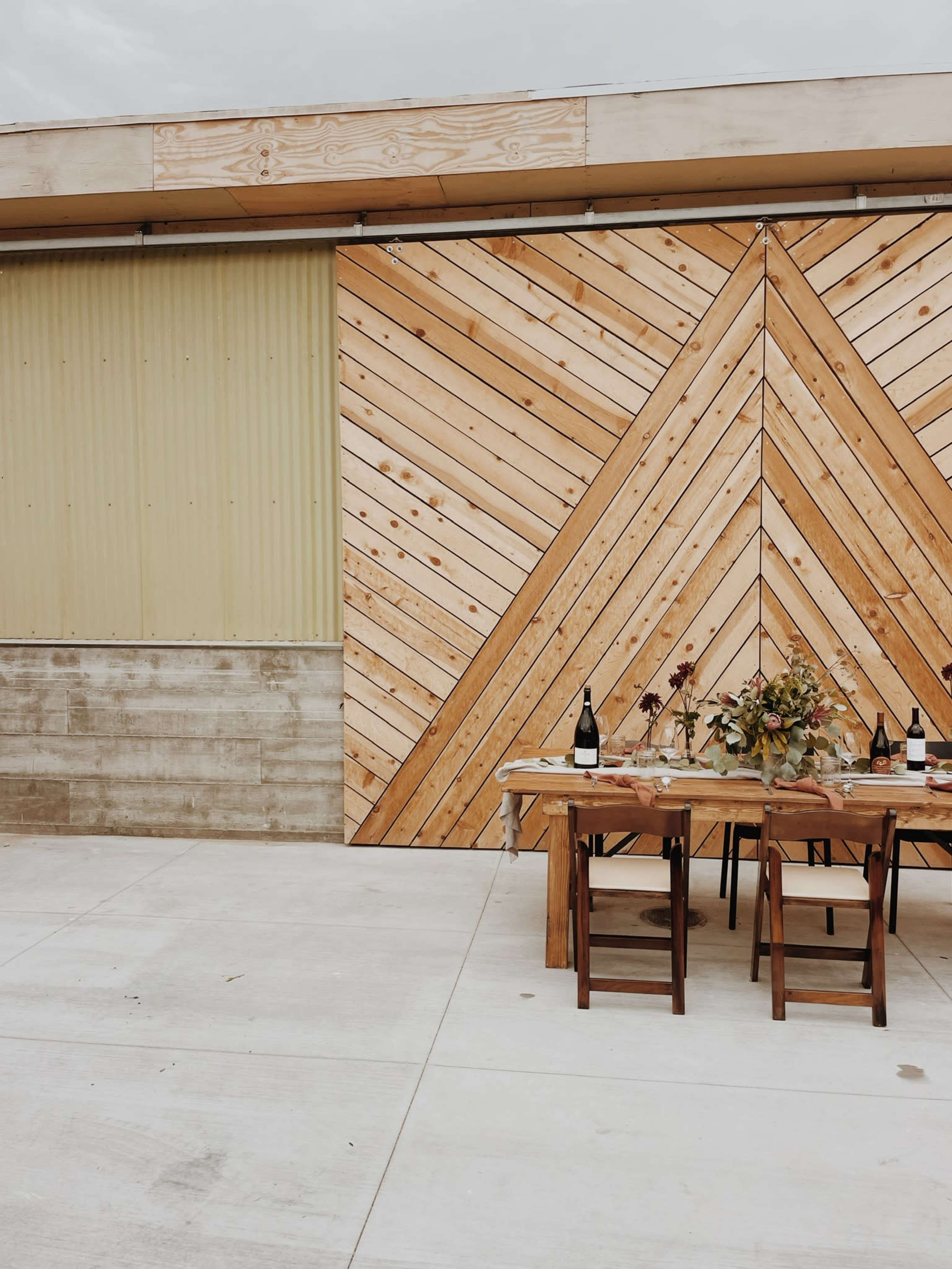 A modern outdoor dining setup with a wooden table and chairs, set against a backdrop of a diagonal wooden panel and a corrugated metal wall.