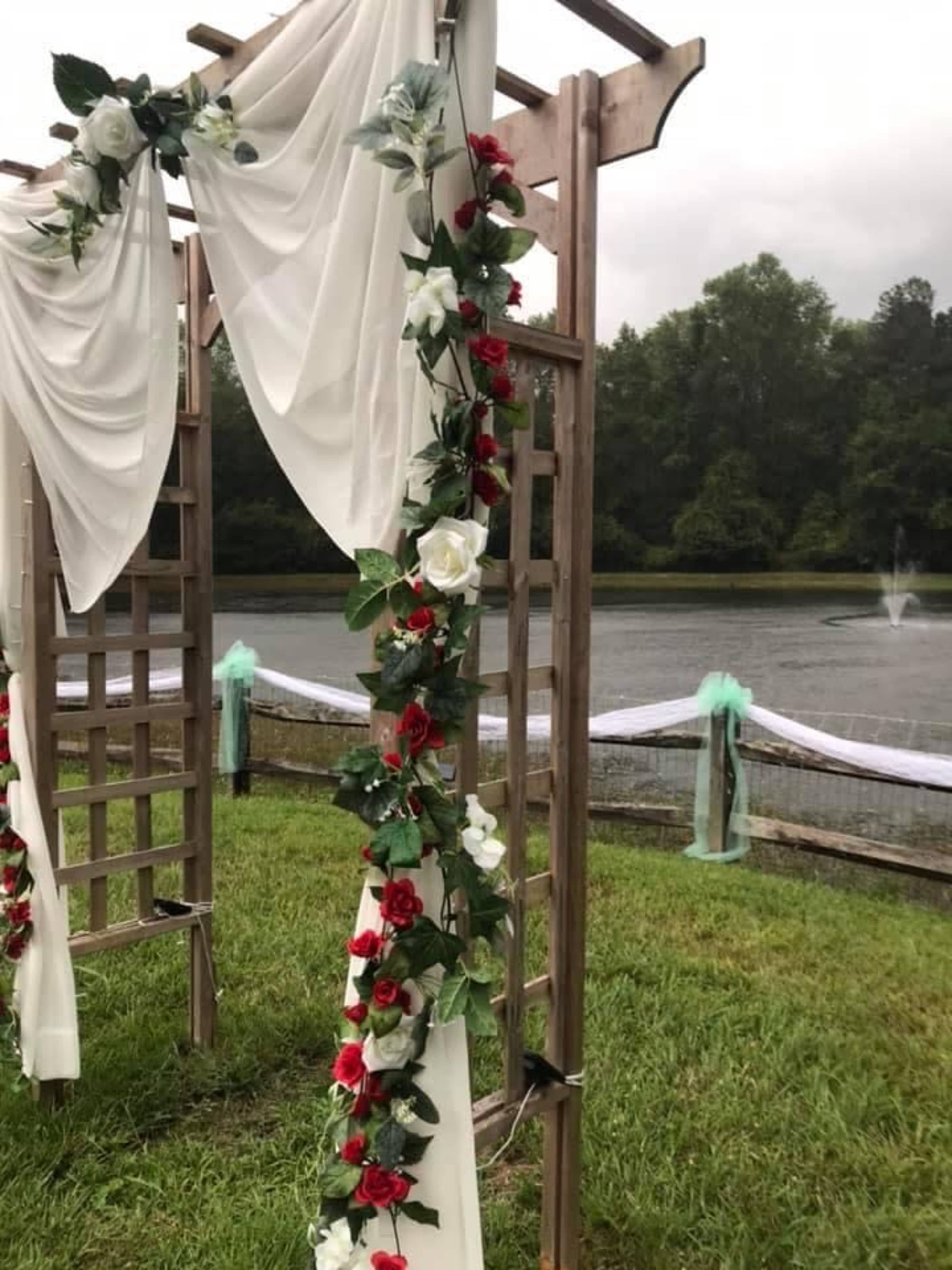 A wooden arch decorated with white fabric and flowers, set by a lake with a fountain in the background.