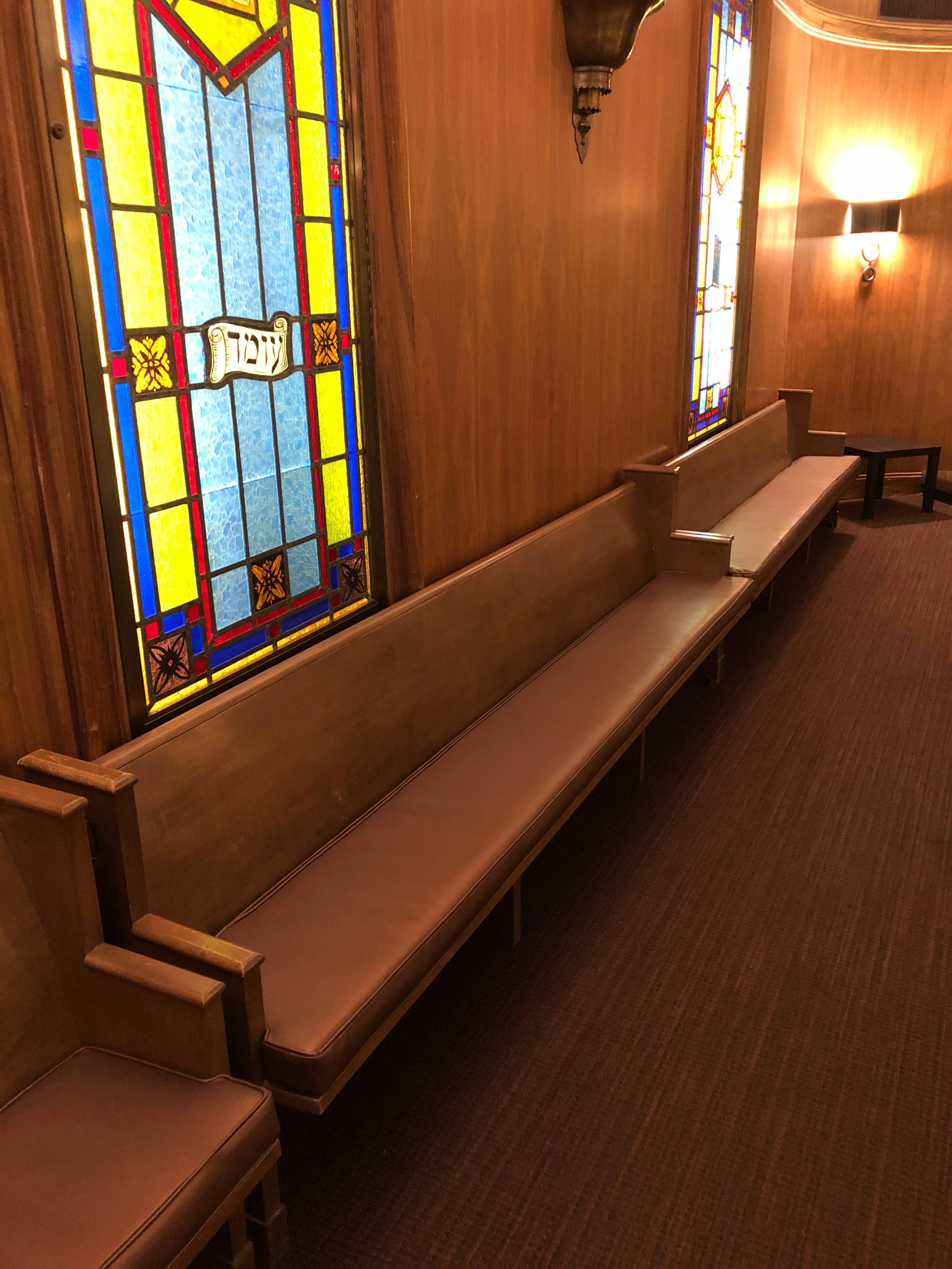 A wooden interior with long benches and stained glass windows on one wall, illuminated by wall sconces.