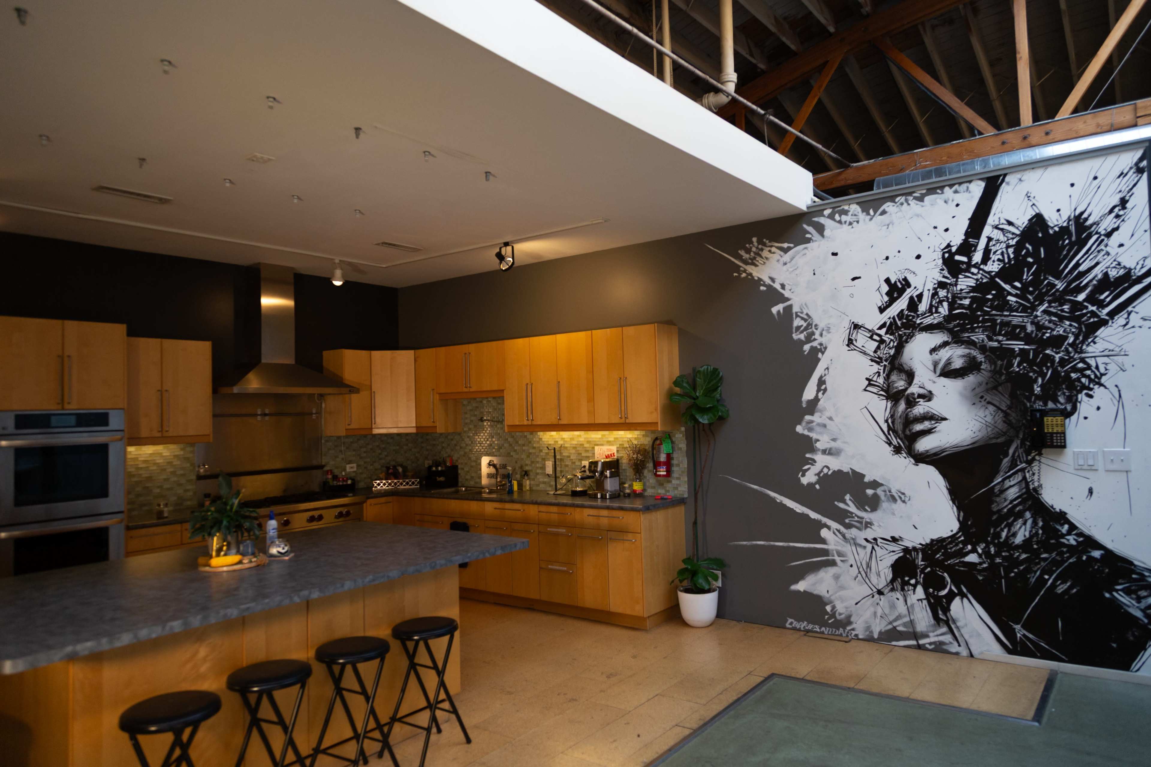 The image shows a modern kitchen with wooden cabinetry and a large mural of a woman on the wall adjacent to it.