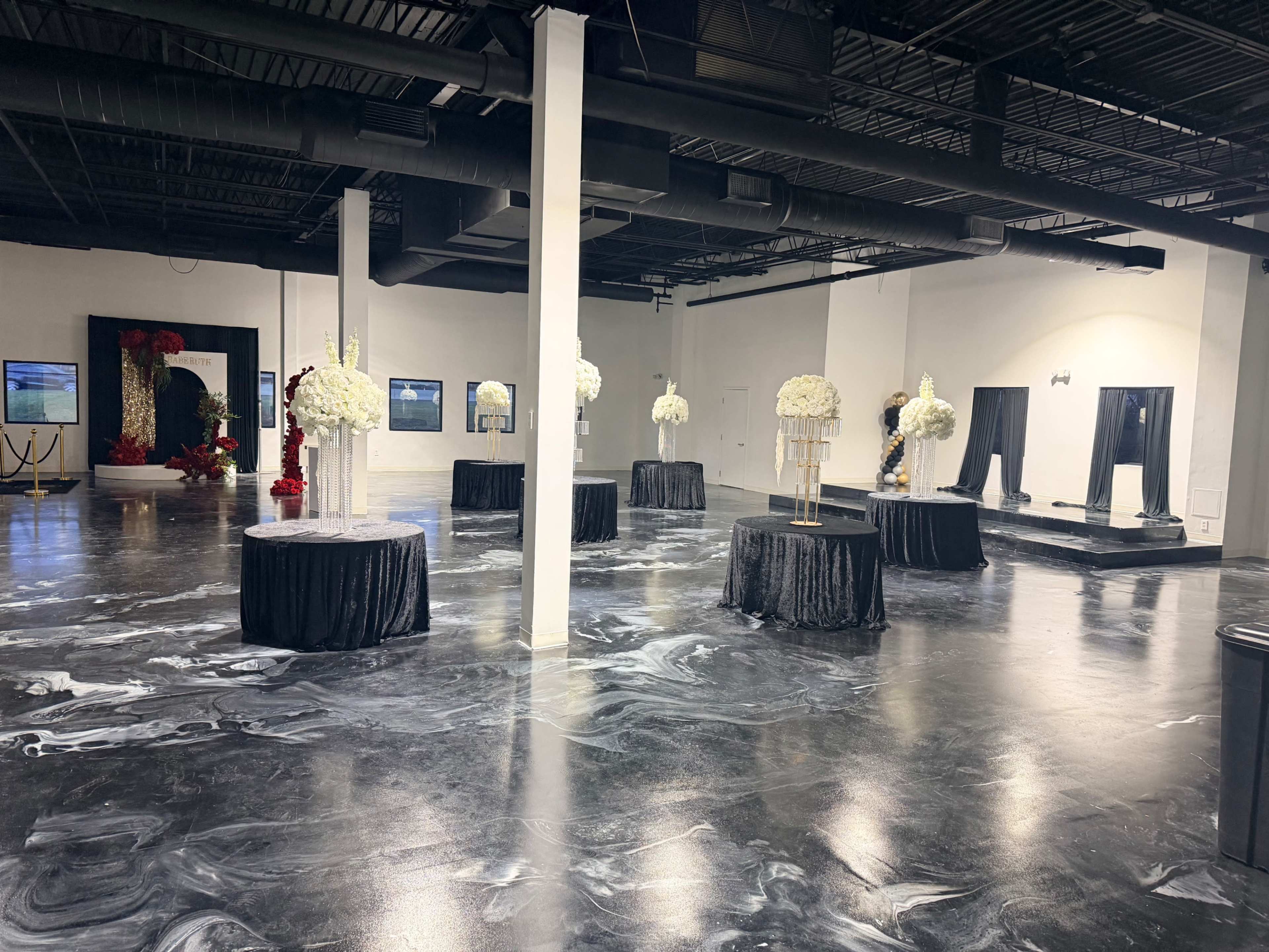 The image shows an event space with several round tables adorned with black tablecloths and floral arrangements, set against a smooth, glossy floor and minimalist decor.