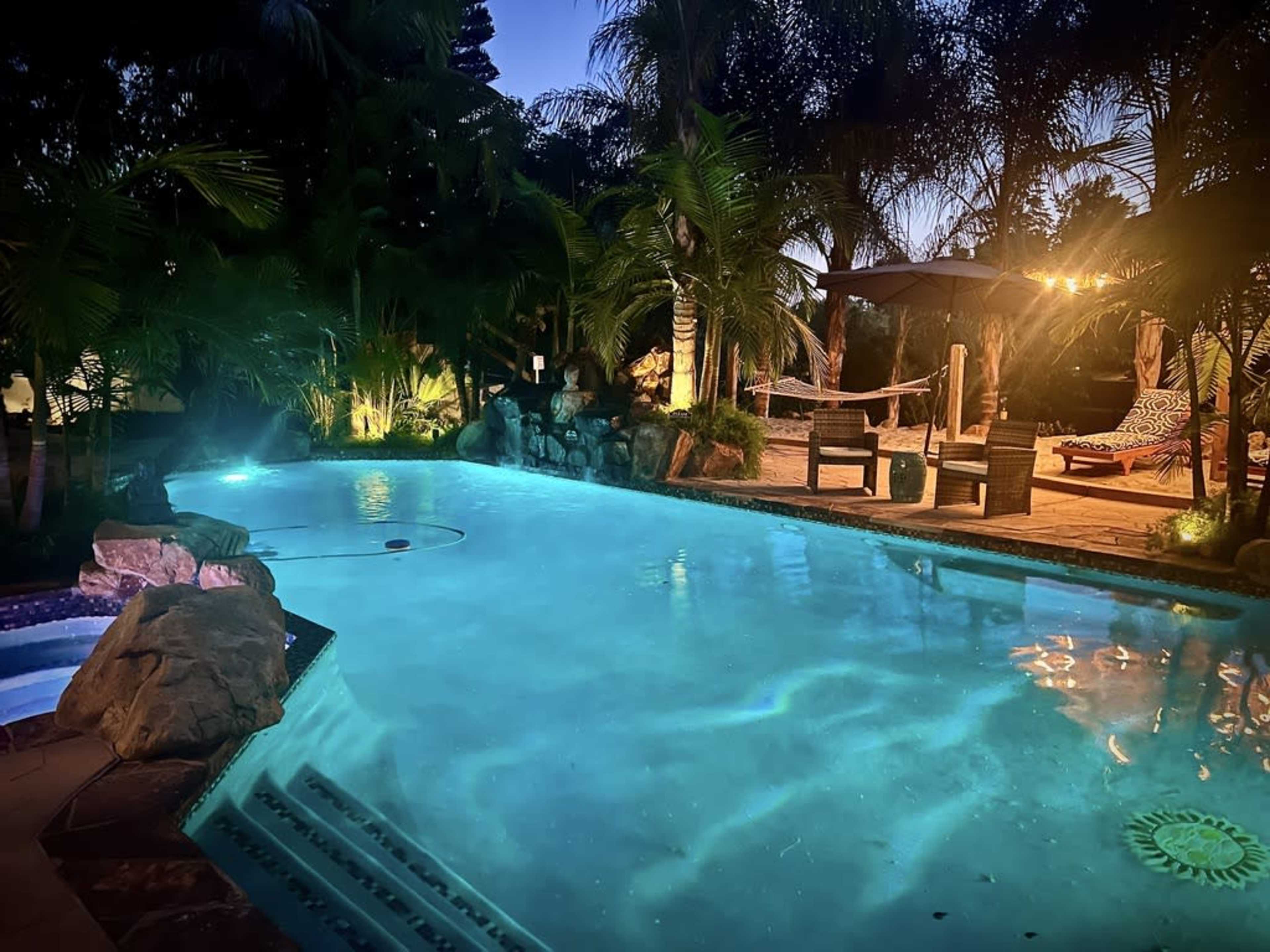 The image shows a lit swimming pool surrounded by palm trees and lounge chairs in a serene outdoor setting at dusk.