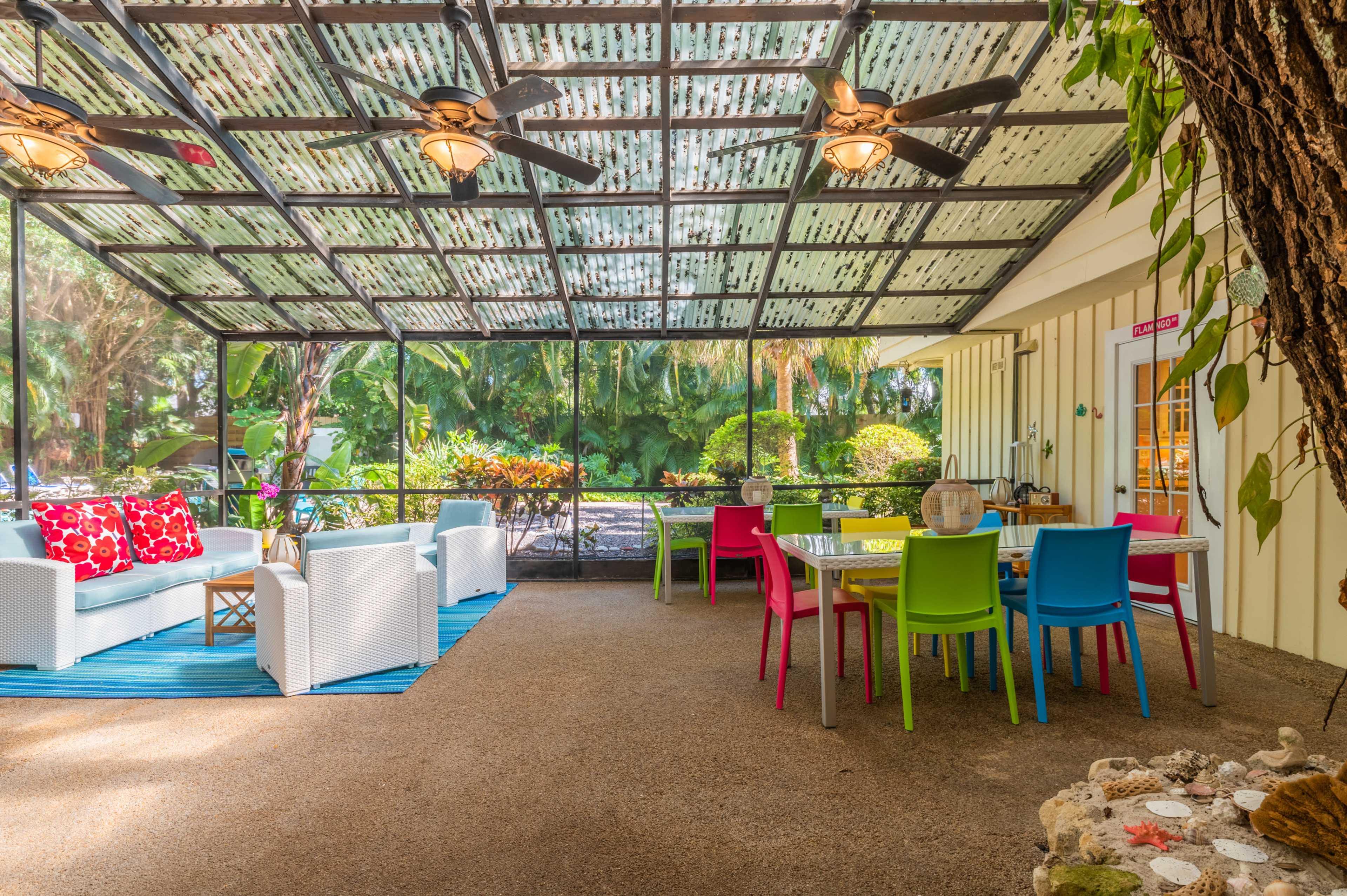 A screened porch featuring colorful chairs and a dining table set within a lush garden setting.