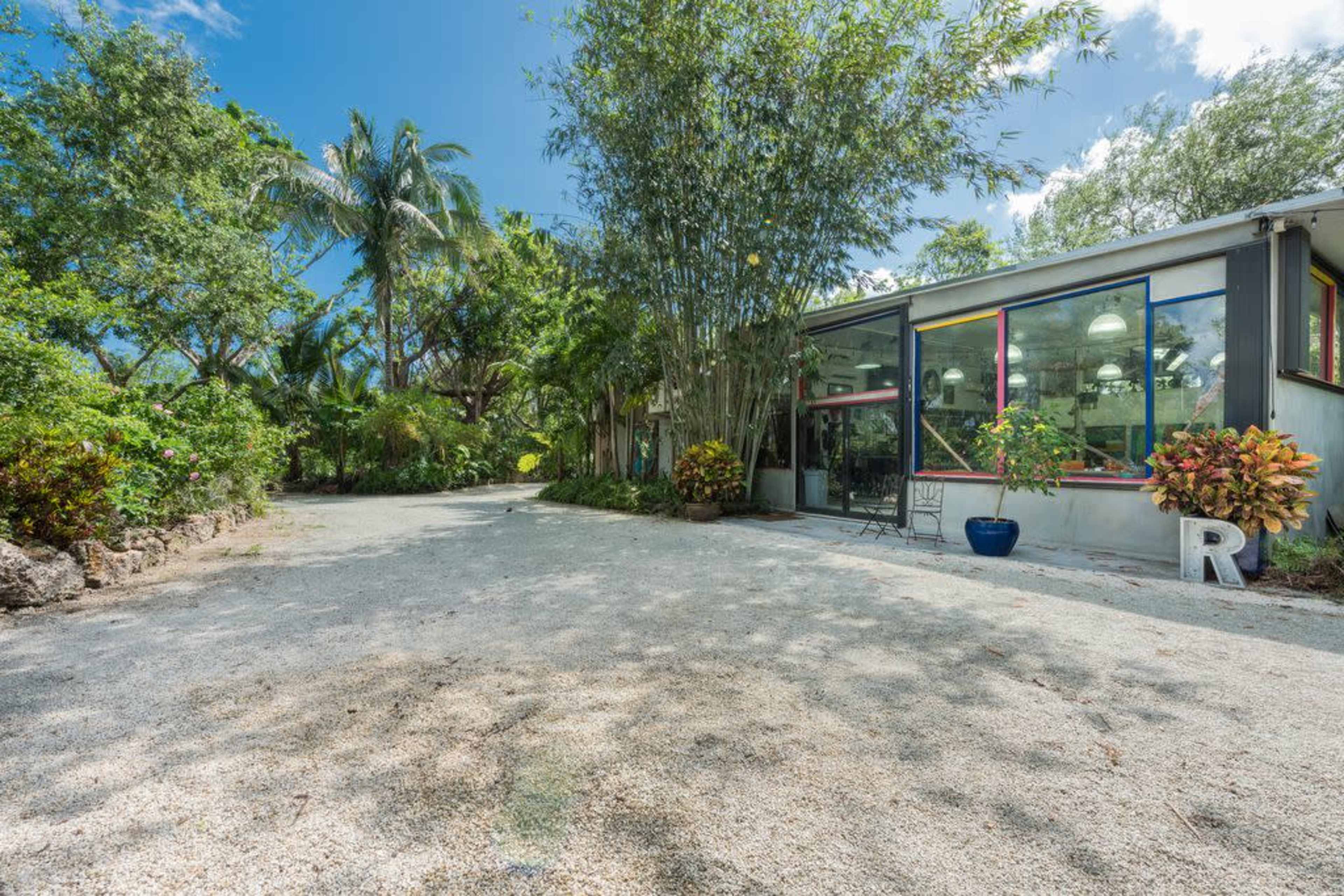 A modern, single-story building with large windows sits amidst lush greenery and palm trees on a gravel pathway.