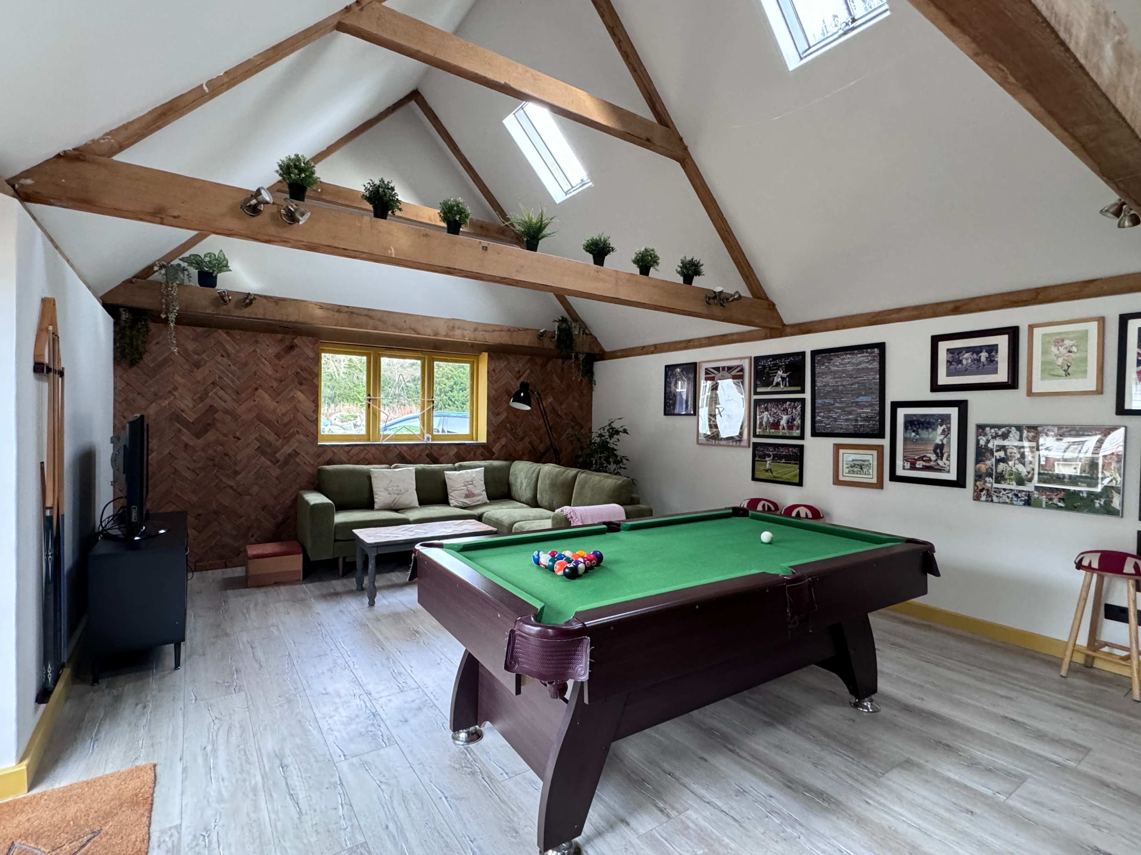 A bright games room features a billiards table, a green sofa, wooden beams, and framed pictures on the walls.