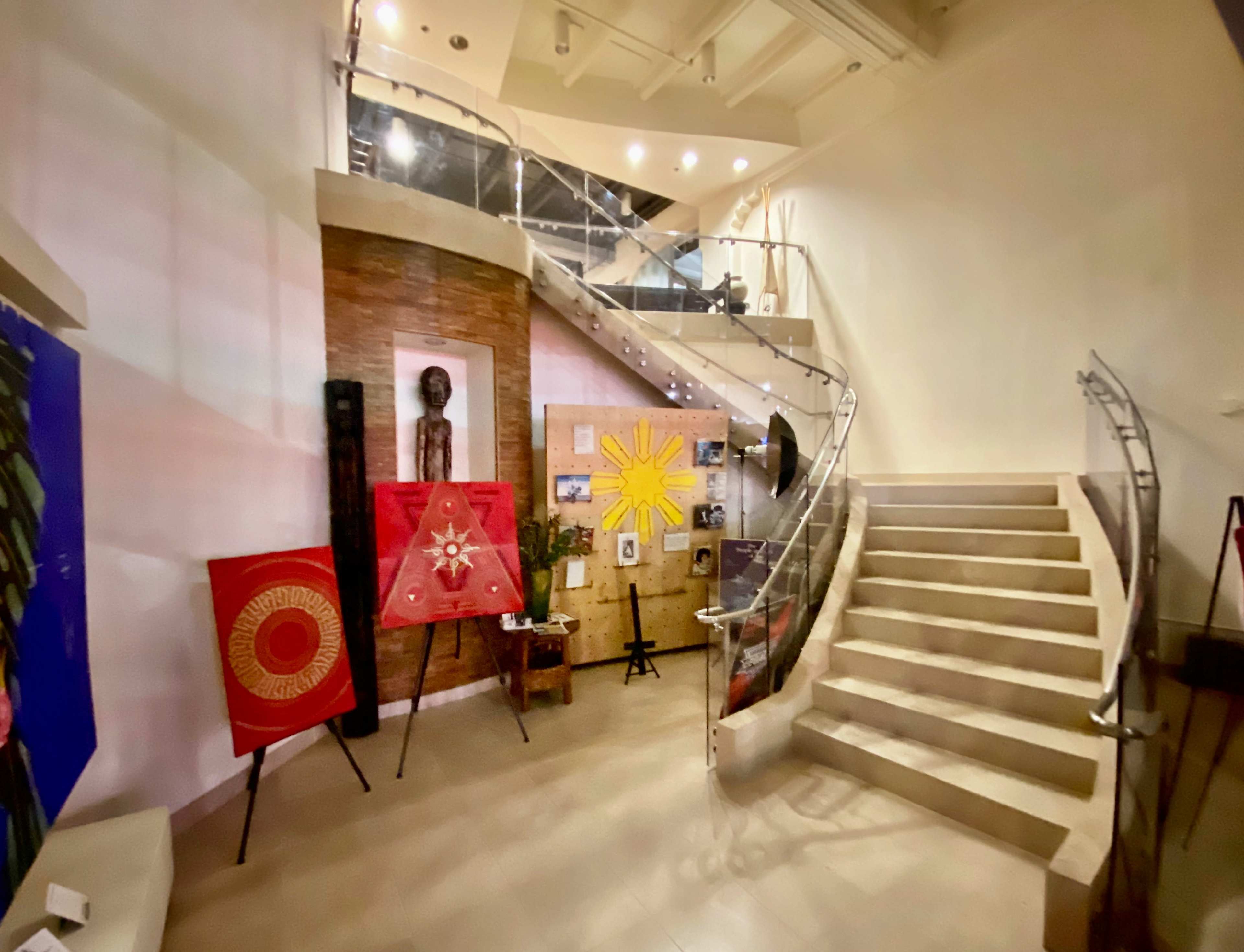 The image shows a modern lobby with a staircase leading to an upper level, featuring artwork on the walls and decorative pieces arranged around the space.
