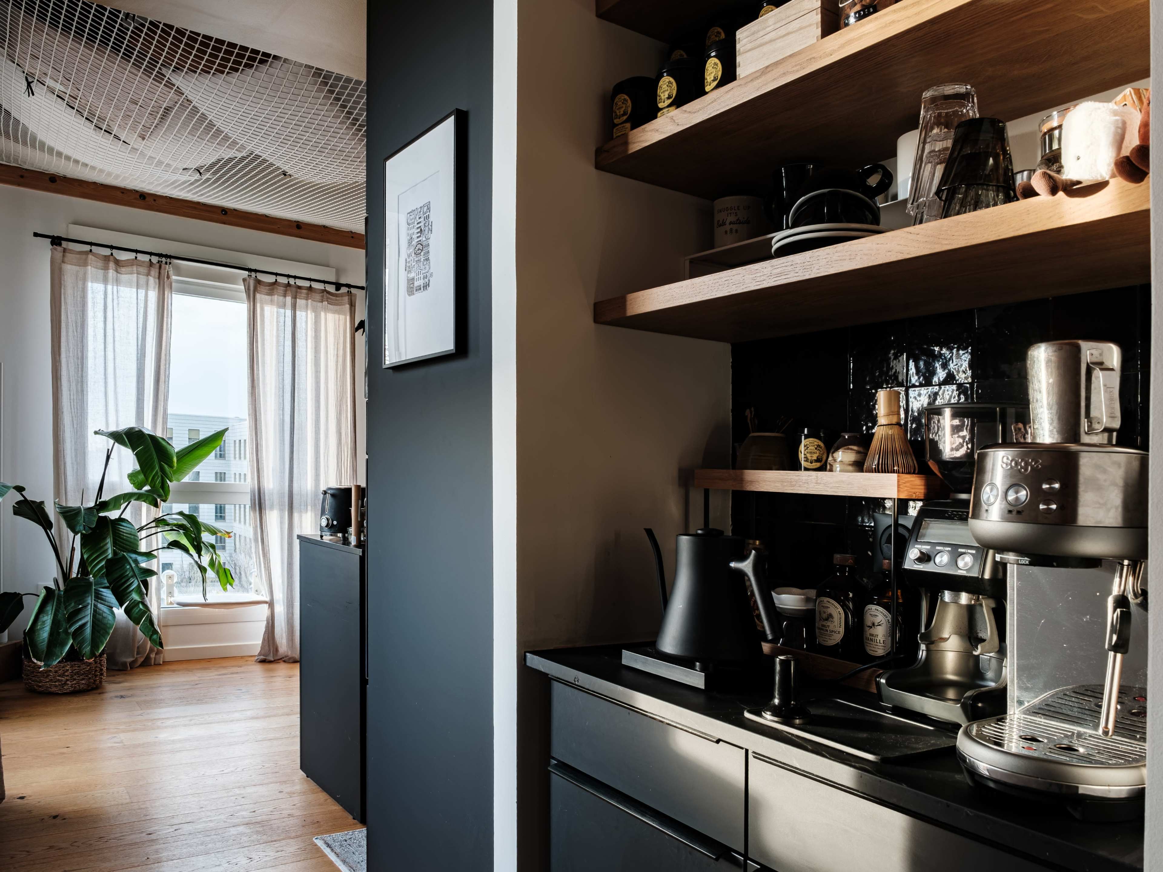 The image shows a modern kitchen area with wooden shelves, an espresso machine, and a large window adorned with light curtains, creating a cozy atmosphere.