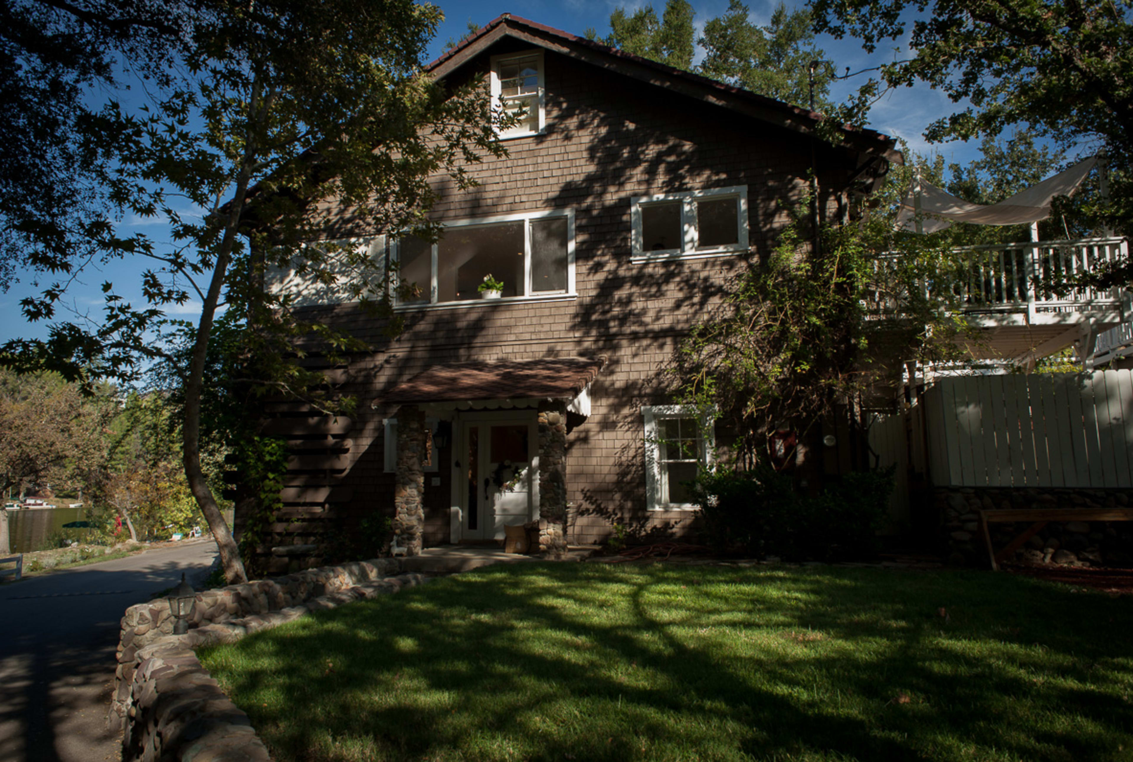 A two-story wooden house with large windows and a garden sits under the shade of trees.